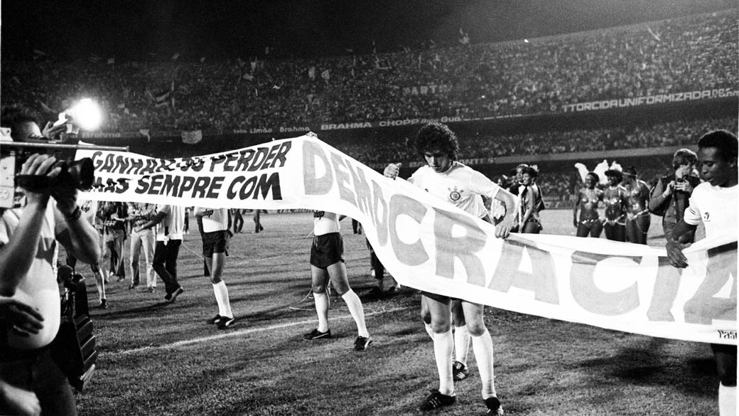 Em 1981, o publicit&aacute;rio e corintiano Washington Olivetto criou a campanha &ldquo;Democracia Corintiana&rdquo;, que apoiou a campanha das Diretas J&aacute;. Acima, jogadores do Corinthians divulgam a campanha em partida contra o S&atilde;o Paulo, v&aacute;lida pela final do Campeonato Paulista de 1983, conquistado pela equipe alvinegra