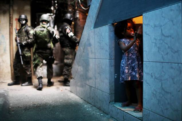 Uma garota é fotografada em uma porta sob uma escada enquanto ao seu redor policiais do Exército patrulham a Favela da Rocinha, durante operação no Rio de janeiro - 25/09/2017 Uma garota é fotografada em uma porta sob uma escada enquanto ao seu redor policiais do Exército patrulham a Favela da Rocinha, durante operação no Rio de janeiro - 25/09/2017