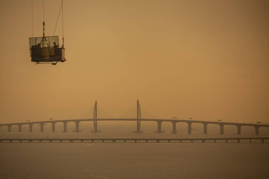 Trabalhadores carregam um painel de vidro em uma plataforma móvel elevada com um trecho da ponte Hong Kong-Zhuhai-Macau ao fundo em Zhuhai, na China - 22/10/2018