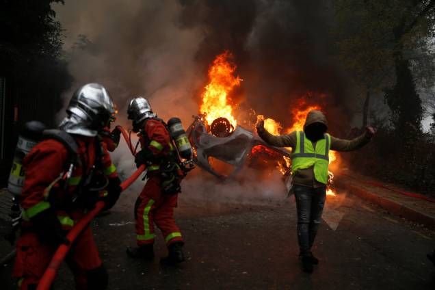 Manifestantes vestindo coletes amarelos, um símbolo de um protesto de motoristas franceses contra impostos mais elevados sobre o diesel, passa perto de um carro em chamas durante os protestos perto da Place de l'Etoile em Paris, França - 01/12/2018 Manifestantes vestindo coletes amarelos, um símbolo de um protesto de motoristas franceses contra impostos mais elevados sobre o diesel, passa perto de um carro em chamas durante os protestos perto da Place de l'Etoile em Paris, França - 01/12/2018