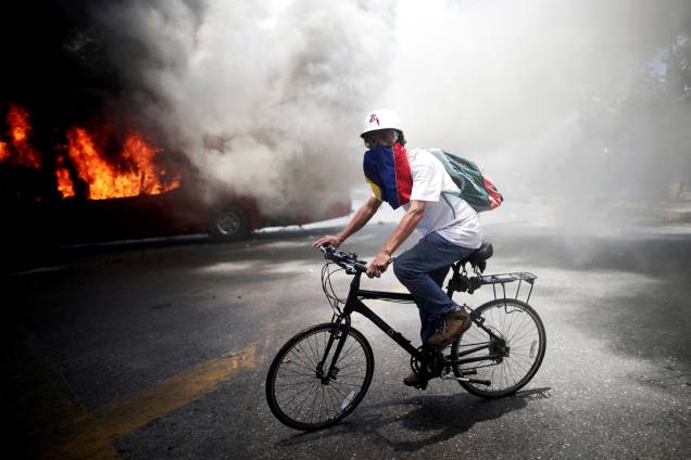 Manifestante da oposição pedala bicicleta próximo de ônibus incendiado, nos arredores da base aérea Generalisimo Francisco de Miranda "La Carlota", em Caracas, capital da Venezuela - 30/04/2019 Manifestante da oposição pedala bicicleta próximo de ônibus incendiado, nos arredores da base aérea Generalisimo Francisco de Miranda "La Carlota", em Caracas, capital da Venezuela - 30/04/2019