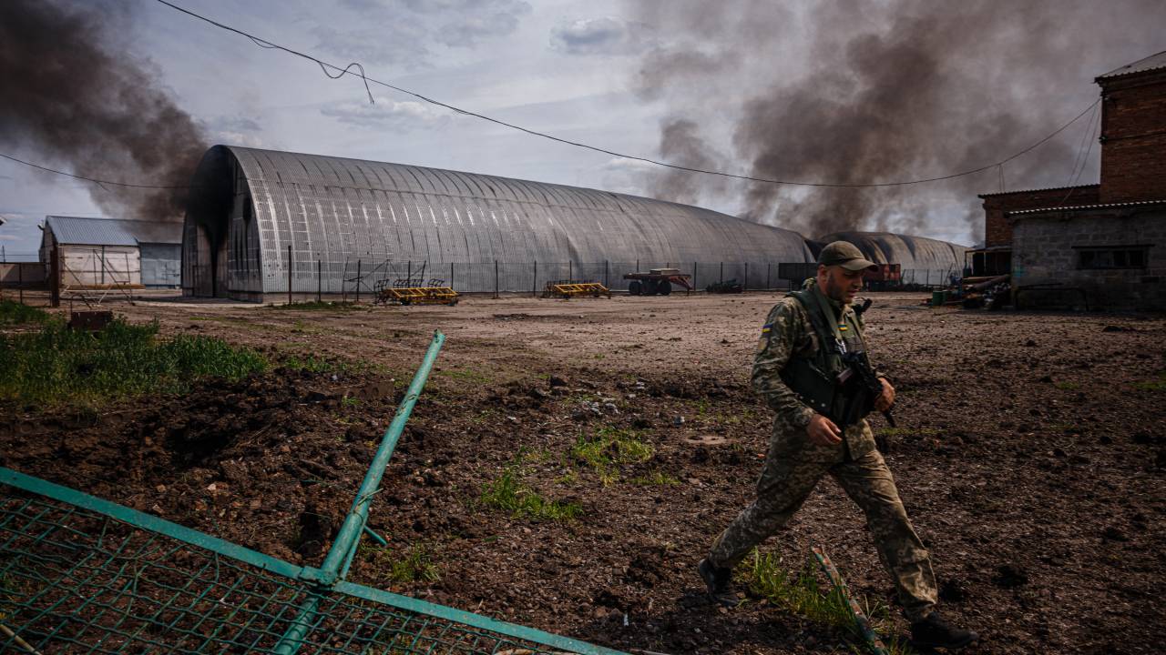 Um membro das Forças de Defesa Territoriais da Ucrânia passa por um prédio de armazenamento em chamas depois de ter sido bombardeado na vila de Temyrivka em 4 de maio de 2022, em meio à invasão russa da Ucrânia. - O incêndio em um prédio de armazenamento do comprimento de um campo de futebol está queimando há pelo menos um dia - mas não há bombeiros em Temyrivka porque todos foram evacuados, deixando a fumaça preta subir sem pressa. Essa aldeia fica em uma terra de ninguém onde os dois lados estão tão próximos, em alguns lugares cerca de 3 kms, que as tropas ucranianas usando binóculos podem ver os russos cavando em suas posições. (Foto de Dimitar DILKOFF/AFP)