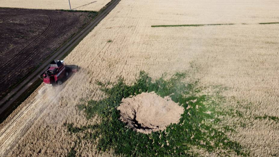 Foto aérea em 07/07/2022, na região Kramatorsk, mostra um agricultor colhendo trigo enquanto dirige uma colheitadeira ao lado uma cratera suspeita de ser causada por um ataque aéreo russo.