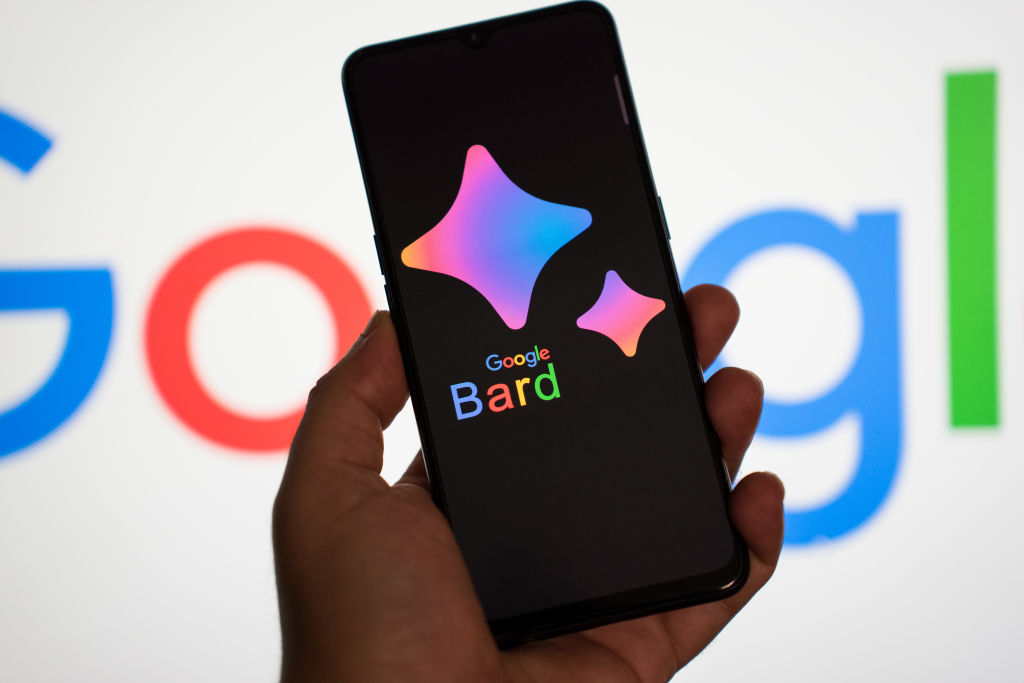 A hand holding a smartphone with the Google Bard logo is seen, in front of a computer screen showing Google's logo. (Photo illustration by Nikos Pekiaridis/NurPhoto via Getty Images)
