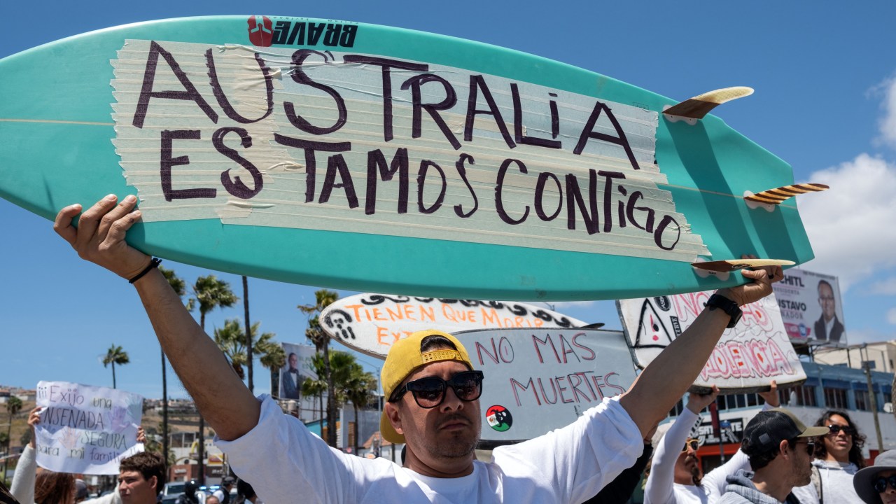 A man raises a surfboard reading "Australia We Are With You" next to members of the surfing community protesting against insecurity after two Australians and an American surfers went missing last week during a surfing trip, in Ensenada, Baja California state, Mexico, on May 5, 2024. Three bodies believed to be those of two Australian brothers and an American who disappeared on a surfing trip in Mexico have bullet wounds to the head, authorities said Sunday. (Photo by Guillermo Arias / AFP)