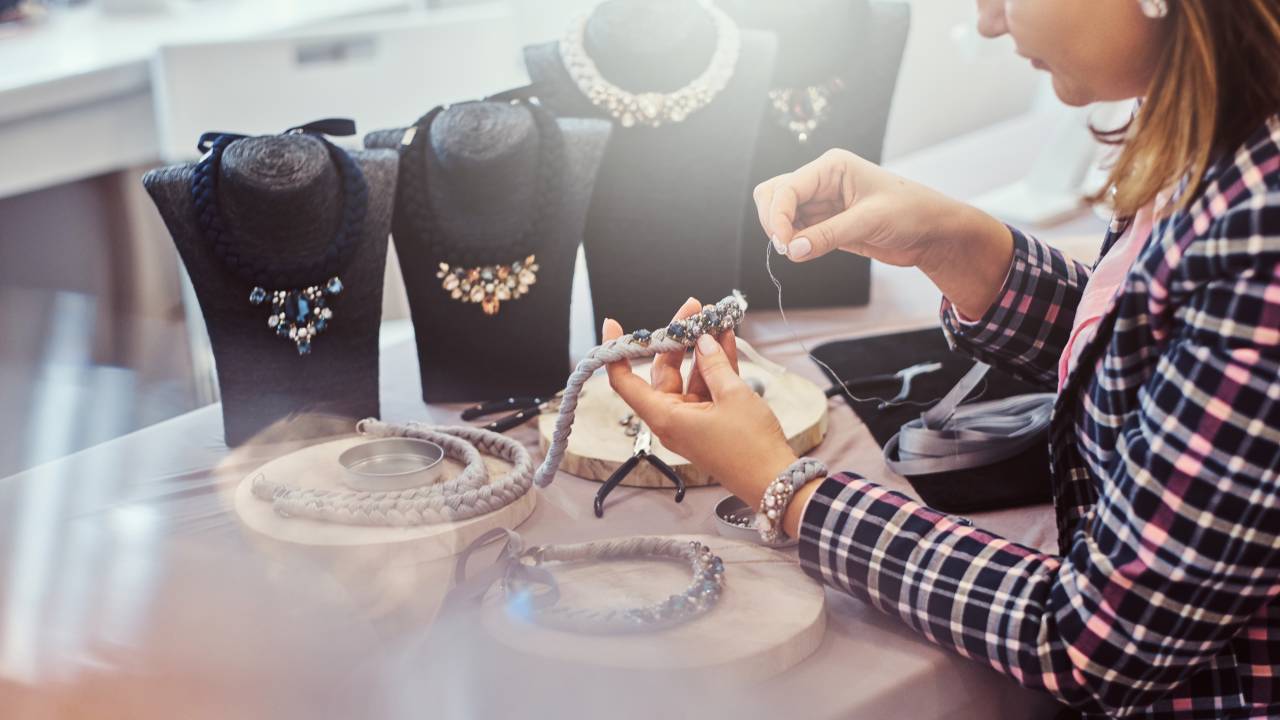 Elegantly dressed woman makes handmade necklaces, working with needles and thread in jewelry workshop.