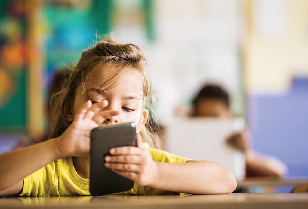 Cute schoolgirl using smart phone on a class at elementary school. Elementary student e-learning over mobile phone during a class in the classroom.
