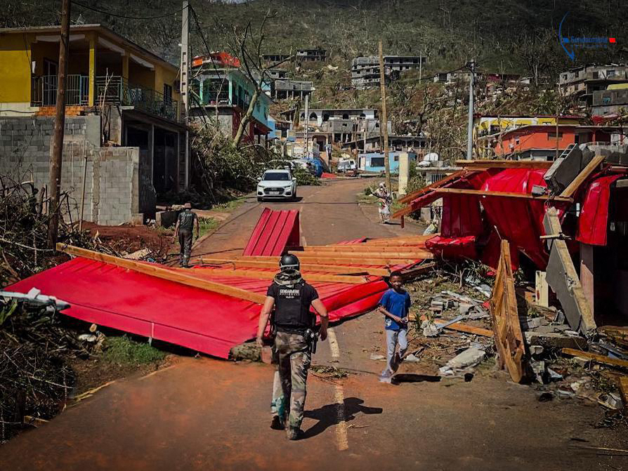 This handout photograph taken on December 15, 2024 and released by the Gendarmerie Nationale on December 16, 2024 shows a boy walking next to a French Gendarme walking along a road blocked by debris during a rescue an emergency operation at an undisclosed location on the French Indian Ocean territory of Mayotte, after the cyclone Chido hit the archipelago. Rescuers raced against time on December 16, 2024 to reach survivors after a devastating cyclone ripped through the French Indian Ocean territory of Mayotte, destroying homes across the islands, with hundreds feared dead. Images from Mayotte, which like other French overseas territories is an integral part of France and ruled from Paris, showed scenes of devastation, with homes reduced to piles of rubble. (Photo by Handout / GENDARMERIE NATIONALE / AFP) / RESTRICTED TO EDITORIAL USE - MANDATORY CREDIT "AFP PHOTO / GENDARMERIE NATIONALE / HANDOUT" - NO MARKETING NO ADVERTISING CAMPAIGNS - DISTRIBUTED AS A SERVICE TO CLIENTS