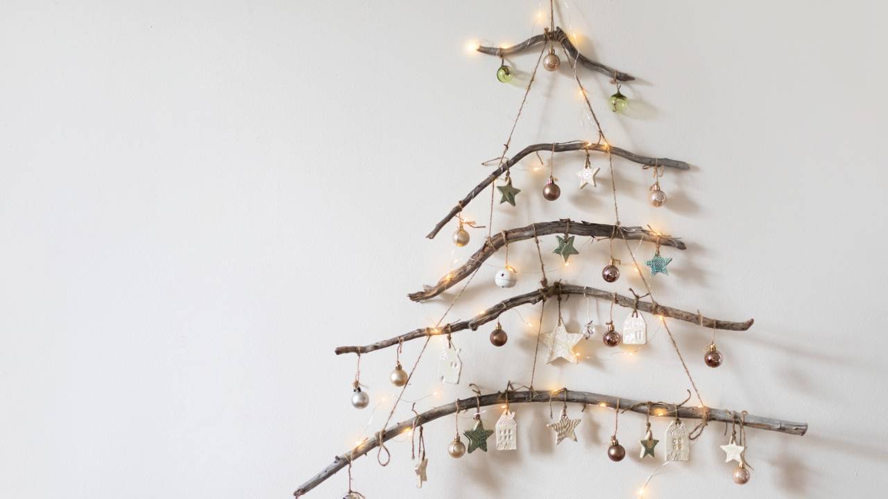 Handmade rustic Christmas tree made from branches with glowing garland lights and ceramic ornaments hanging on white wall. DIY inpiration. Christmas and New Year concept.