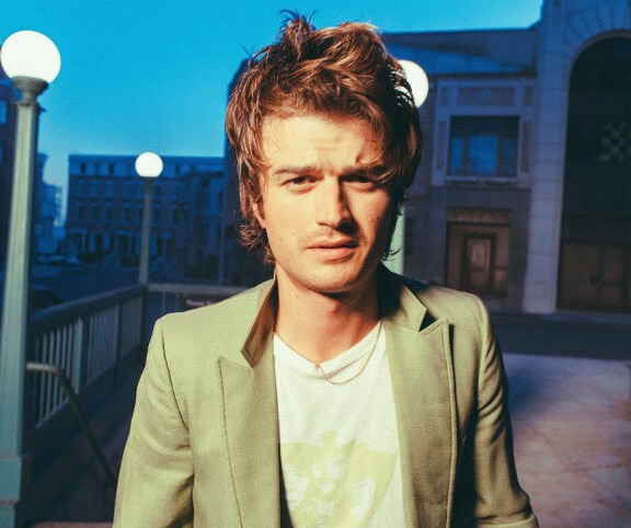 O ator e cantor Joe Keery com uma expressão séria em meio a uma cidade de fundo azulado