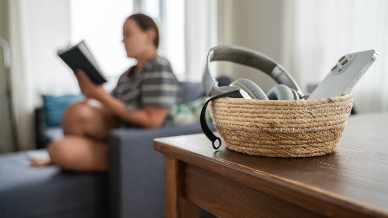 Digital and information detox without devices to promote mindfulness and reduce screen time. Woman reading on background. Basket with smartphone, smartwatch and headphones