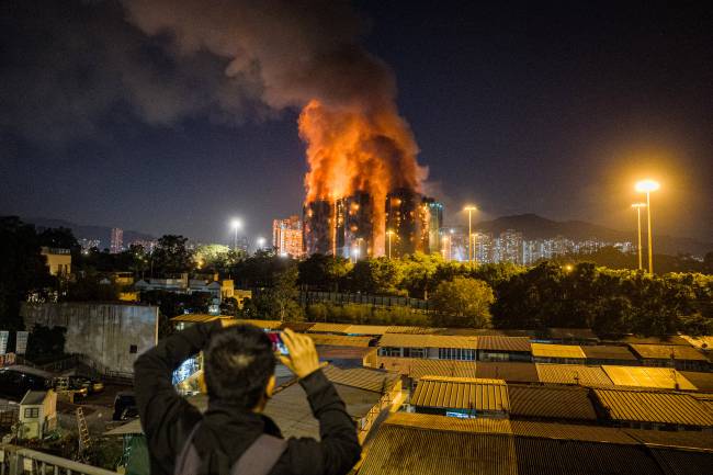An onlooker takes photos as a major fire engulfs several apartment blocks at the Wang Fuk Court residential estate (background) in Hong Kong's Tai Po district on November 26, 2025. At least four people were killed when a fire engulfed several high-rise blocks in a Hong Kong residential estate on November 26, the government said, with media reporting that some residents were trapped inside. (Photo by AFP)