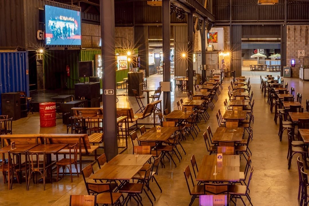 Interior de um grande sal&atilde;o com mesas e cadeiras de madeira escuras, dispostas em longas filas. H&aacute; um tel&atilde;o exibindo uma imagem azulada no canto superior esquerdo, e barris vermelhos no ch&atilde;o. O ambiente tem ilumina&ccedil;&atilde;o quente e industrial, com pilares escuros e estruturas met&aacute;licas aparentes