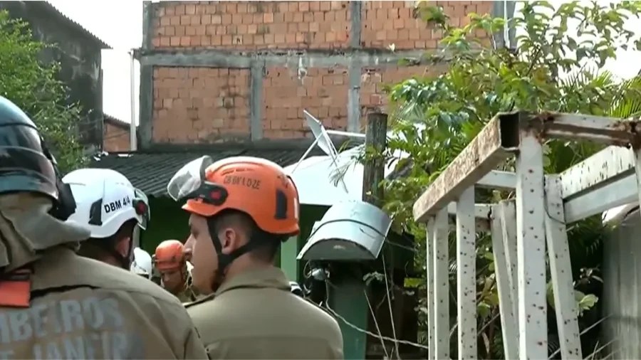 Bombeiros com capacetes de proteção, um laranja e um branco, em área urbana com casas de tijolos e vegetação, possivelmente em operação de resgate ou vistoria