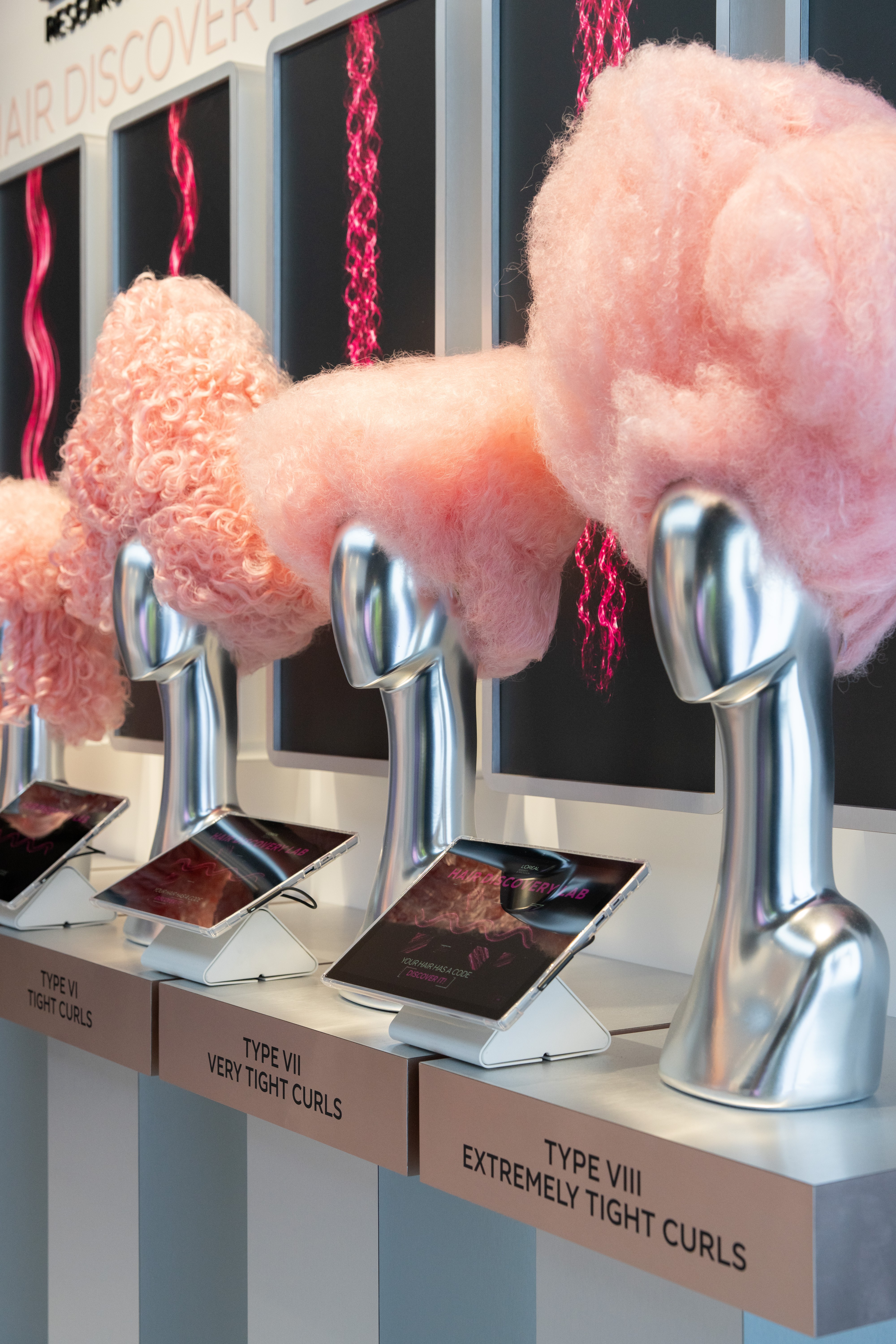 Exposi&ccedil;&atilde;o de tipos de cachos com manequins prateados exibindo perucas de algod&atilde;o doce rosa e telas mostrando padr&otilde;es de cabelo em neon rosa, com tablets e legendas TYPE VI TIGHT CURLS, TYPE VII VERY TIGHT CURLS e TYPE VIII EXTREMELY TIGHT CURLS