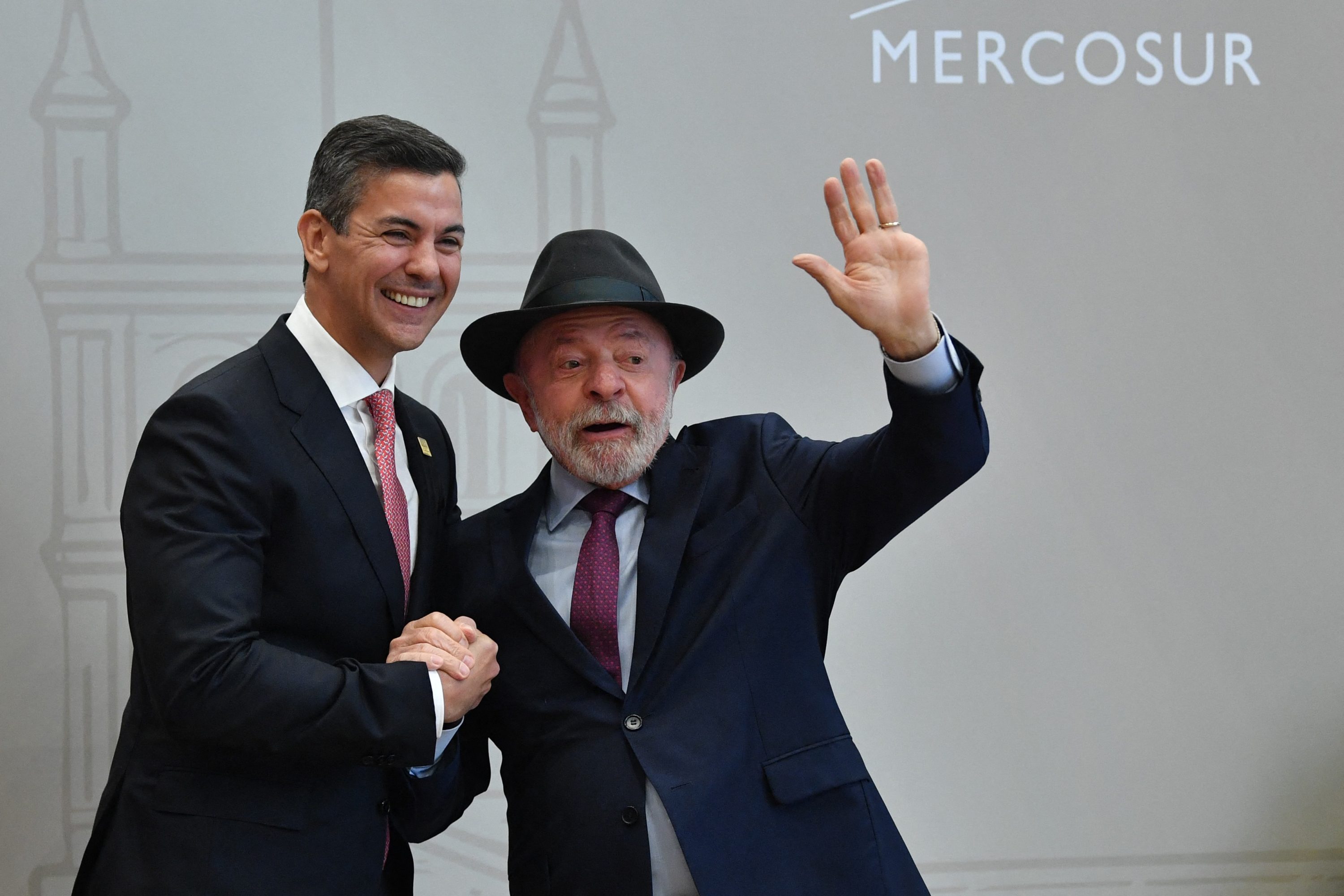Dois homens em ternos escuros, um sorrindo e outro acenando com a mão direita, em frente a uma parede clara com o texto MERCOSUL e um desenho arquitetônico