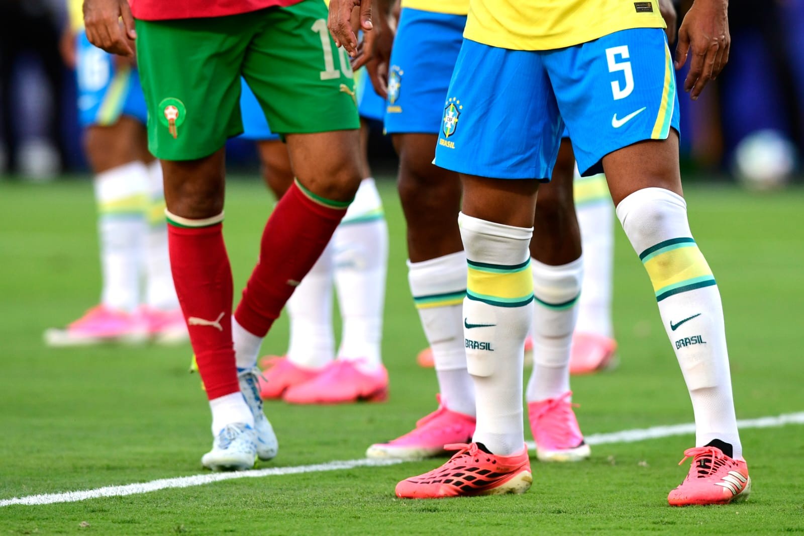 Pernas de jogadores de futebol em campo verde, um com shorts e meias vermelhas, outro com shorts azuis e meias brancas com faixas amarelas e verdes, e chuteiras coloridas