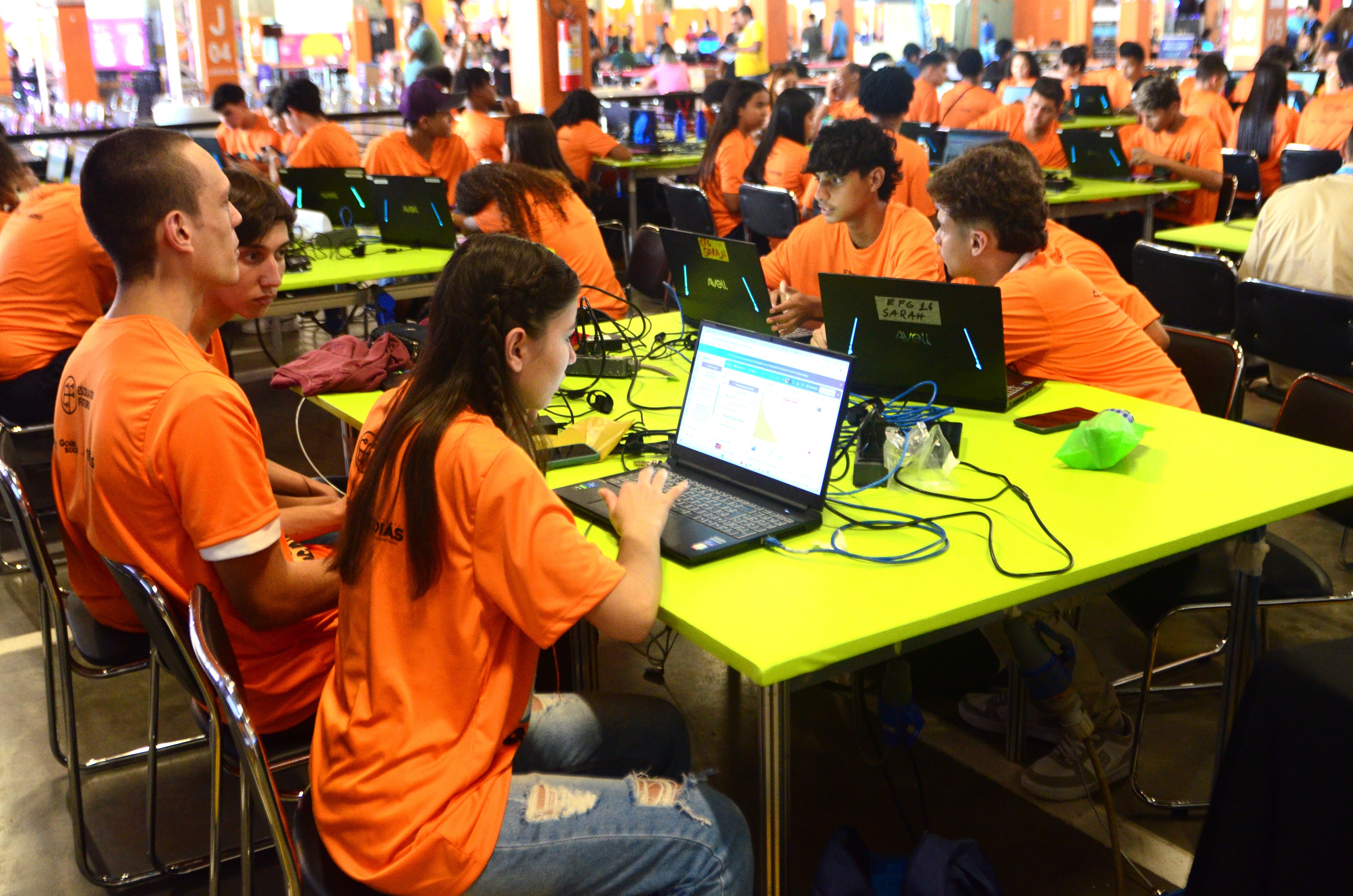 Jovens e adultos, a maioria vestindo camisetas laranja, participam de um evento de tecnologia, sentados em mesas verdes com notebooks e fios espalhados, concentrados nas telas