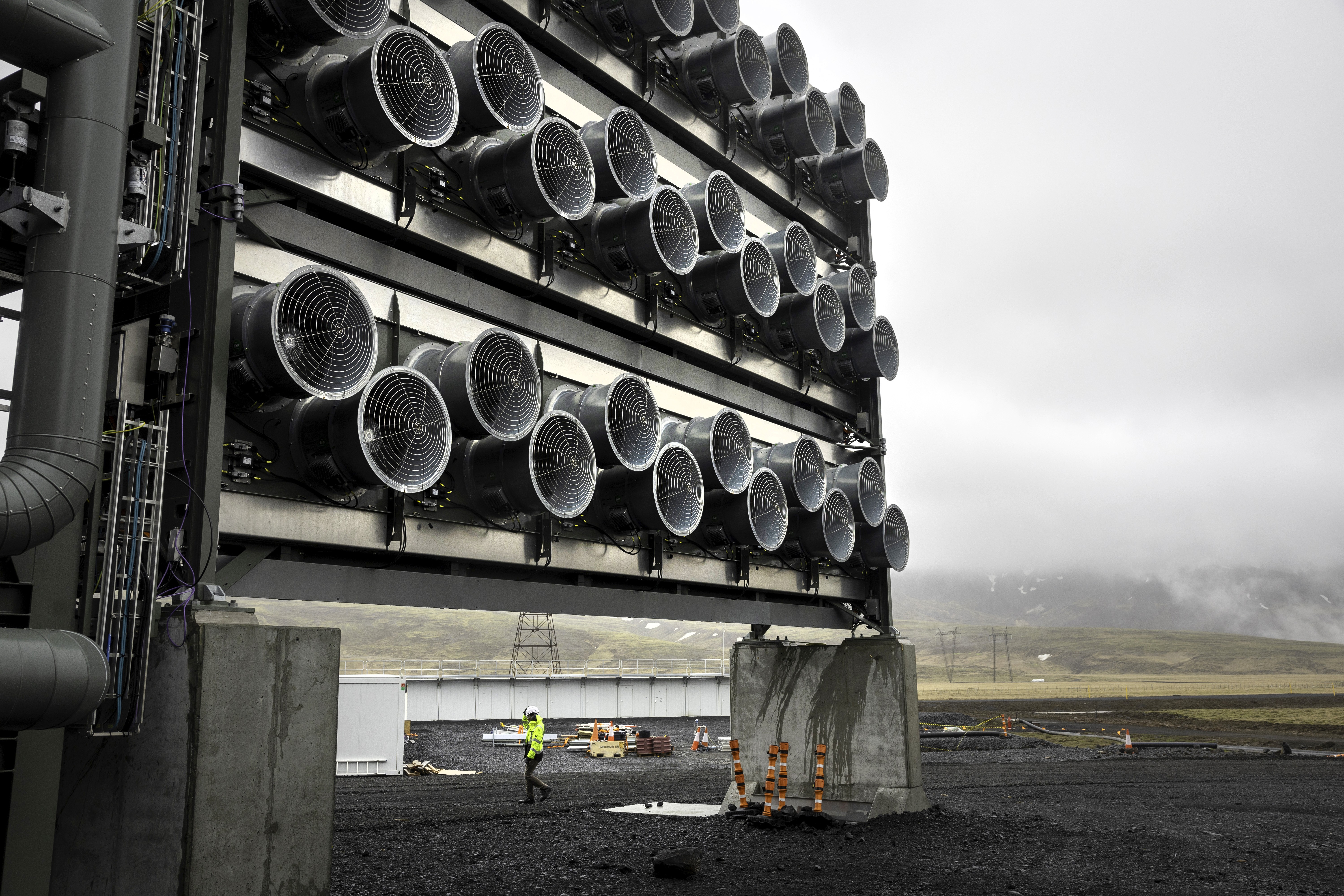 Estrutura industrial com dezenas de grandes ventiladores cinzas em fileiras, um trabalhador de colete verde e capacete branco caminha no chão escuro, com montanhas enevoadas ao fundo