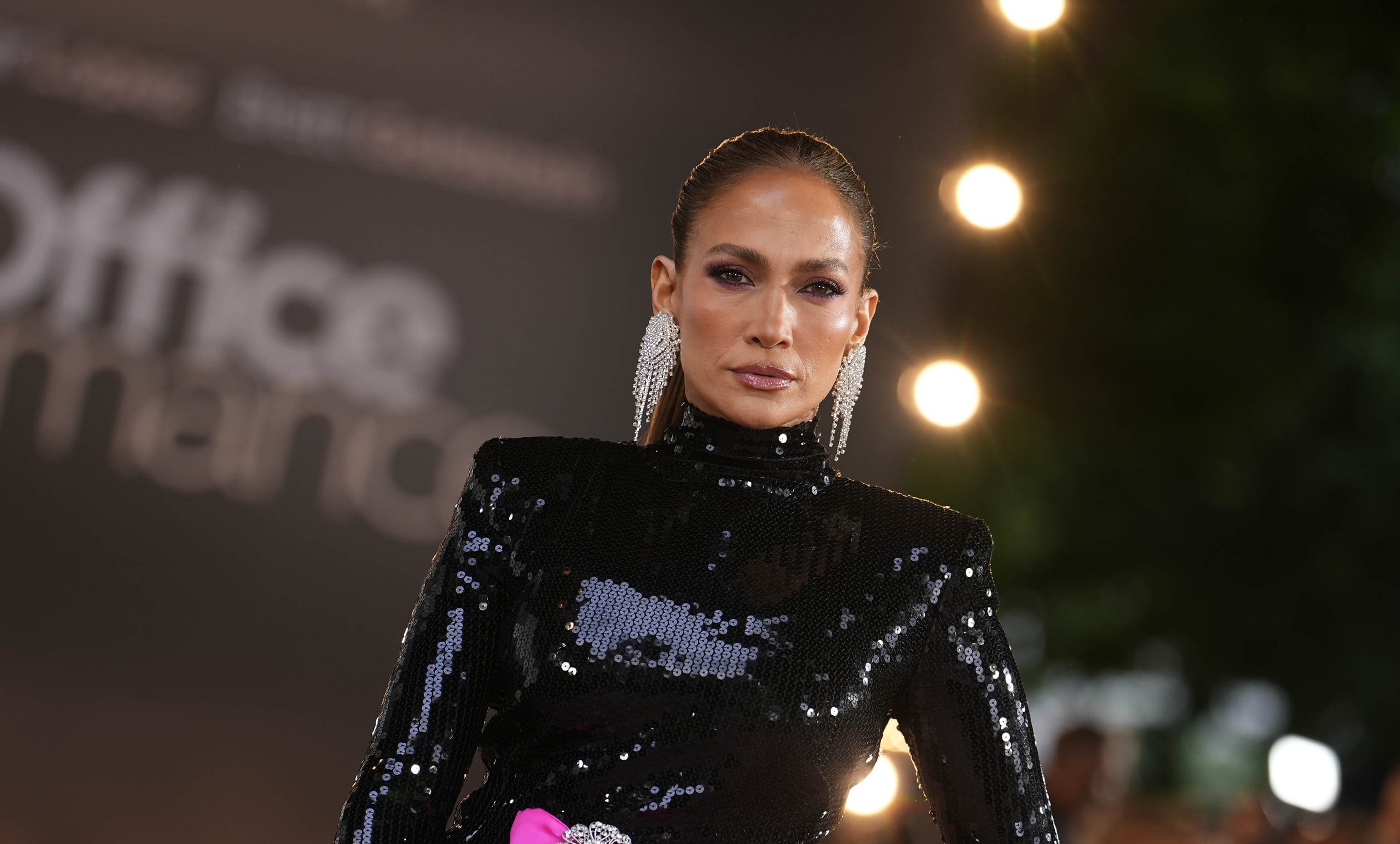 Jennifer Lopez, com cabelo preso e maquiagem marcante, usa um vestido preto de gola alta e lantejoulas, com ombreiras e brincos longos prateados. Ao fundo, luzes desfocadas e parte da palavra Office em branco.