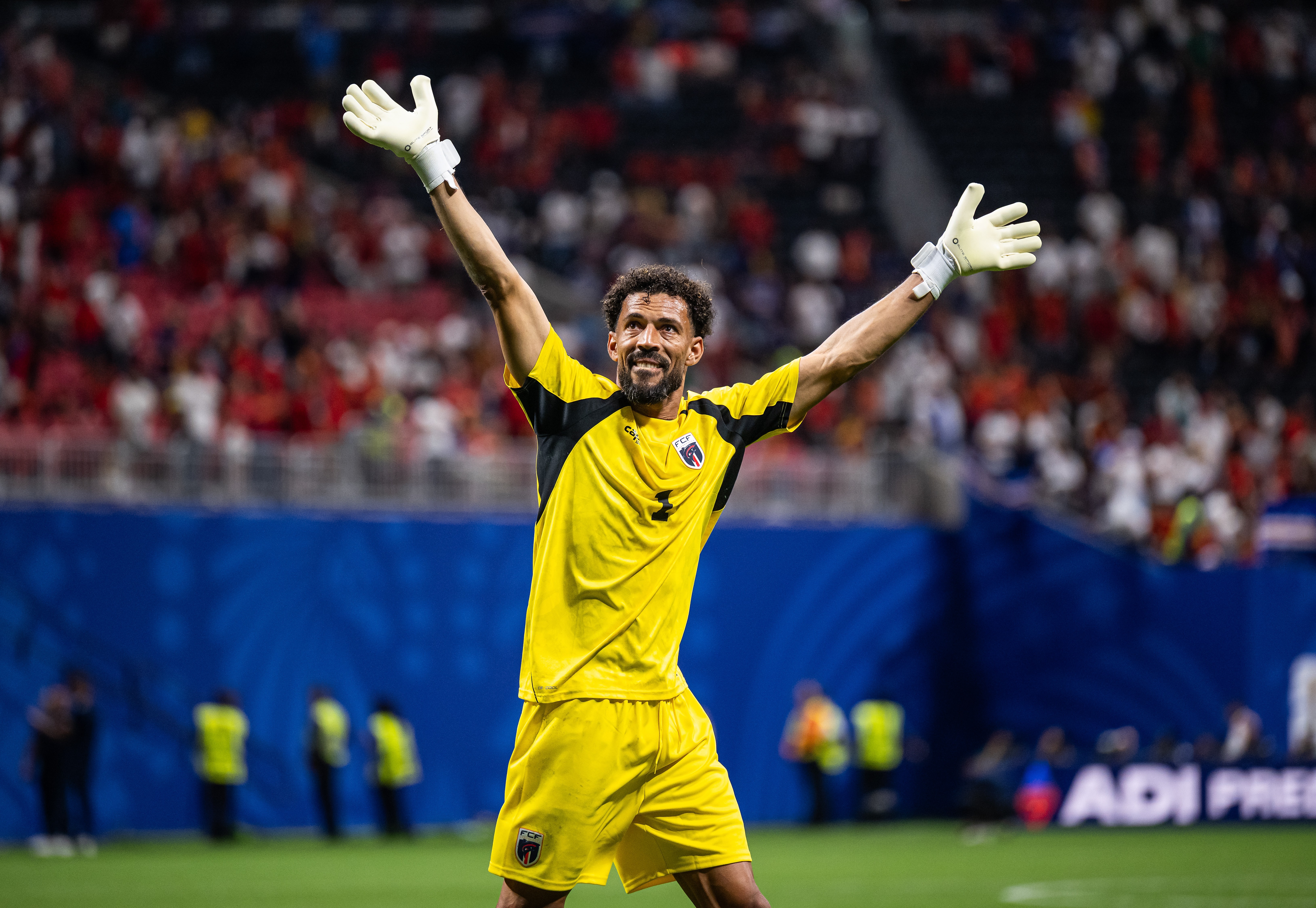 Goleiro de pele morena e barba, vestindo uniforme amarelo e luvas brancas, sorri com os braços erguidos em comemoração no campo de futebol. Torcida e placas de publicidade desfocadas ao fundo