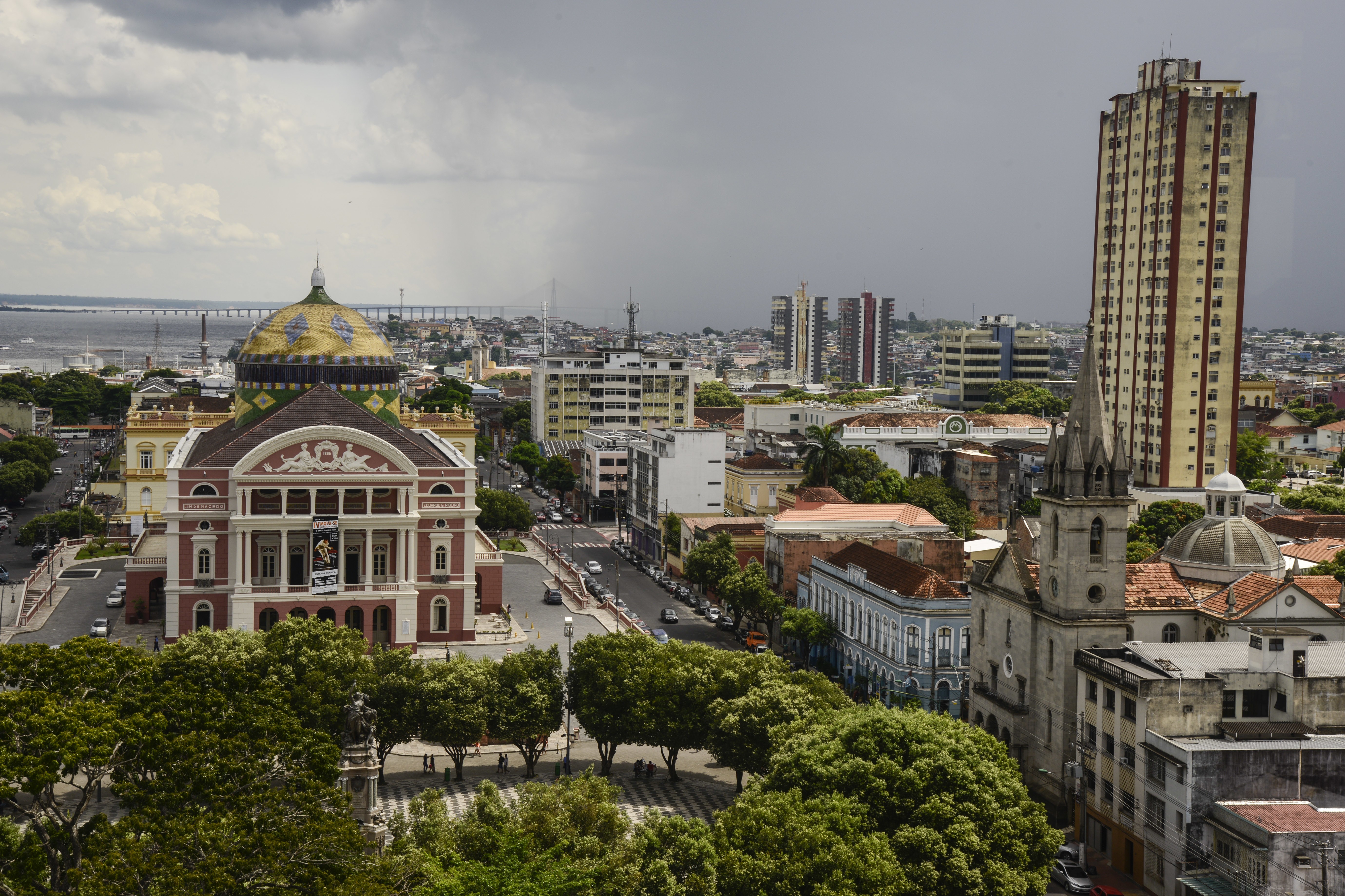 Vista aérea de Manaus com o Teatro Amazonas em primeiro plano, destacando sua cúpula dourada e verde. Ao fundo, prédios altos, uma igreja com torre pontiaguda e a Ponte Rio Negro sob céu nublado. Árvores verdes preenchem a parte inferior da imagem, com ruas e carros visíveis.
