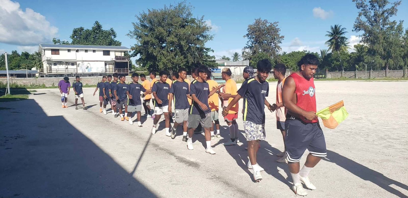 Grupo de jovens atletas caminhando em fila por um campo de terra batida, com um pr&eacute;dio branco e &aacute;rvores ao fundo, sob c&eacute;u azul. Um deles, &agrave; direita, veste camiseta vermelha e segura uma bandeira de &aacute;rbitro de futebol