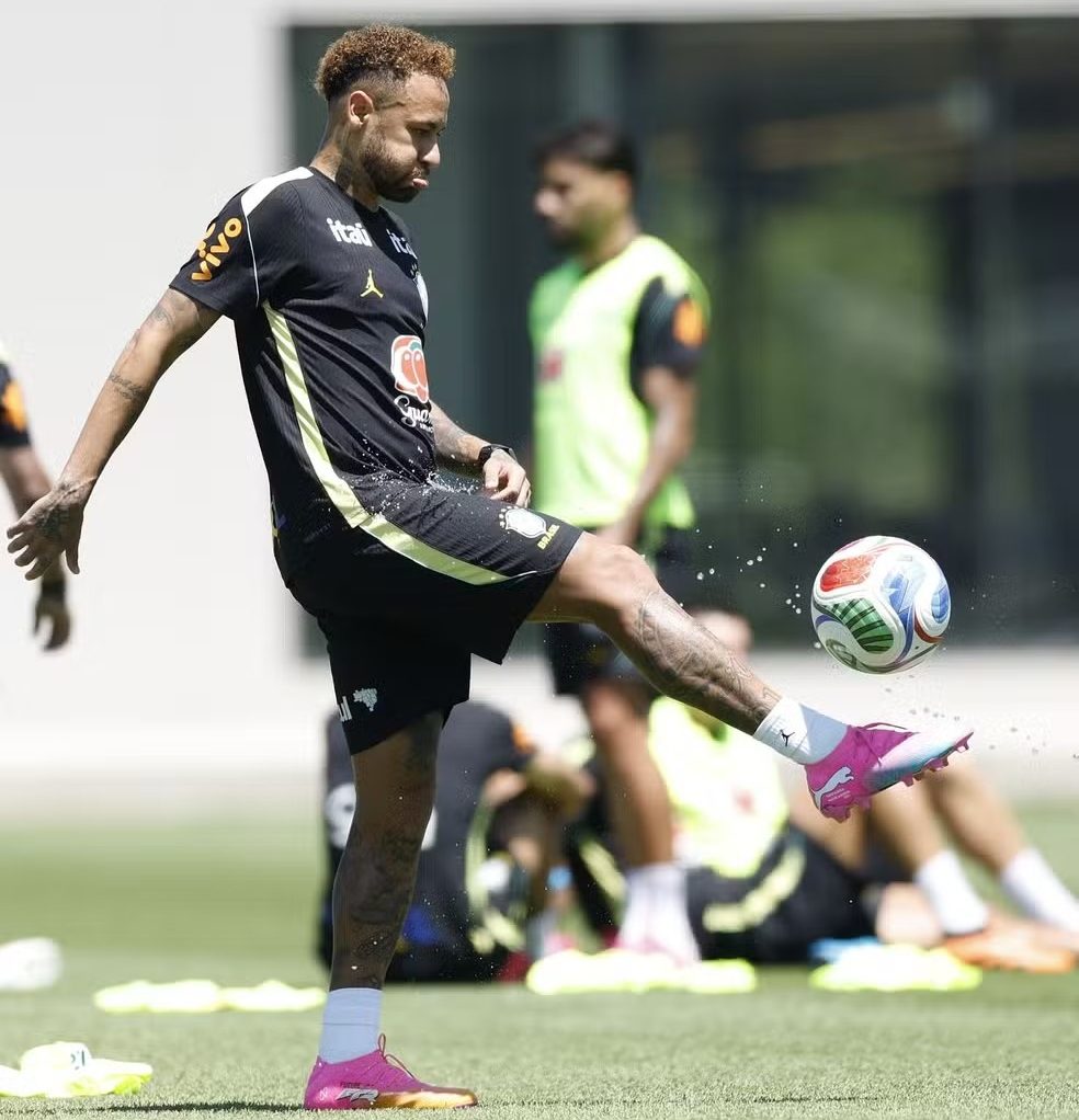 Neymar, com cabelo loiro e barba, chuta uma bola de futebol colorida em campo verde, usando uniforme preto e chuteiras rosa. Got&iacute;culas de &aacute;gua voam ao redor da bola. Outros jogadores desfocados est&atilde;o ao fundo
