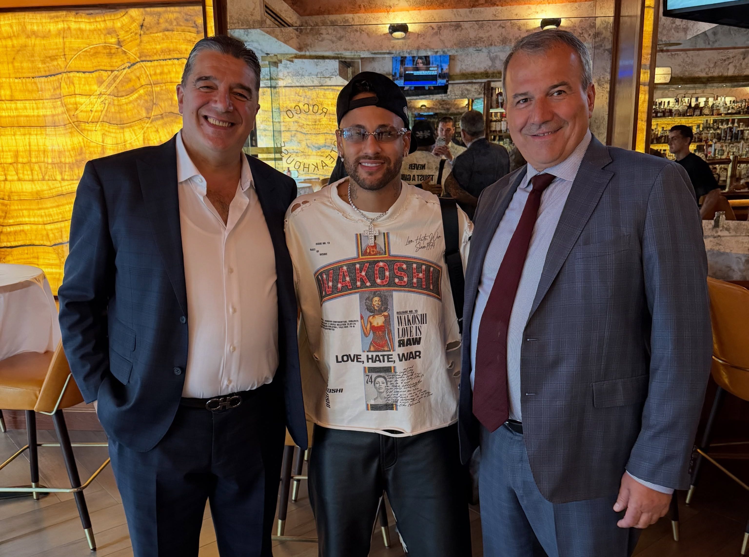 Tr&ecirc;s homens sorridentes posam em um restaurante. &Agrave; esquerda, um homem de terno azul marinho e camisa branca. No centro, Neymar Jr. com bon&eacute; preto, &oacute;culos, camiseta branca estampada e bermuda de couro. &Agrave; direita, outro homem de terno cinza e gravata vinho.
