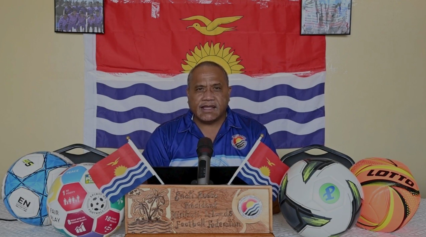 Homem de pele escura, camisa polo azul, falando ao microfone, com a bandeira de Kiribati ao fundo e bolas de futebol e bandeiras pequenas na frente
