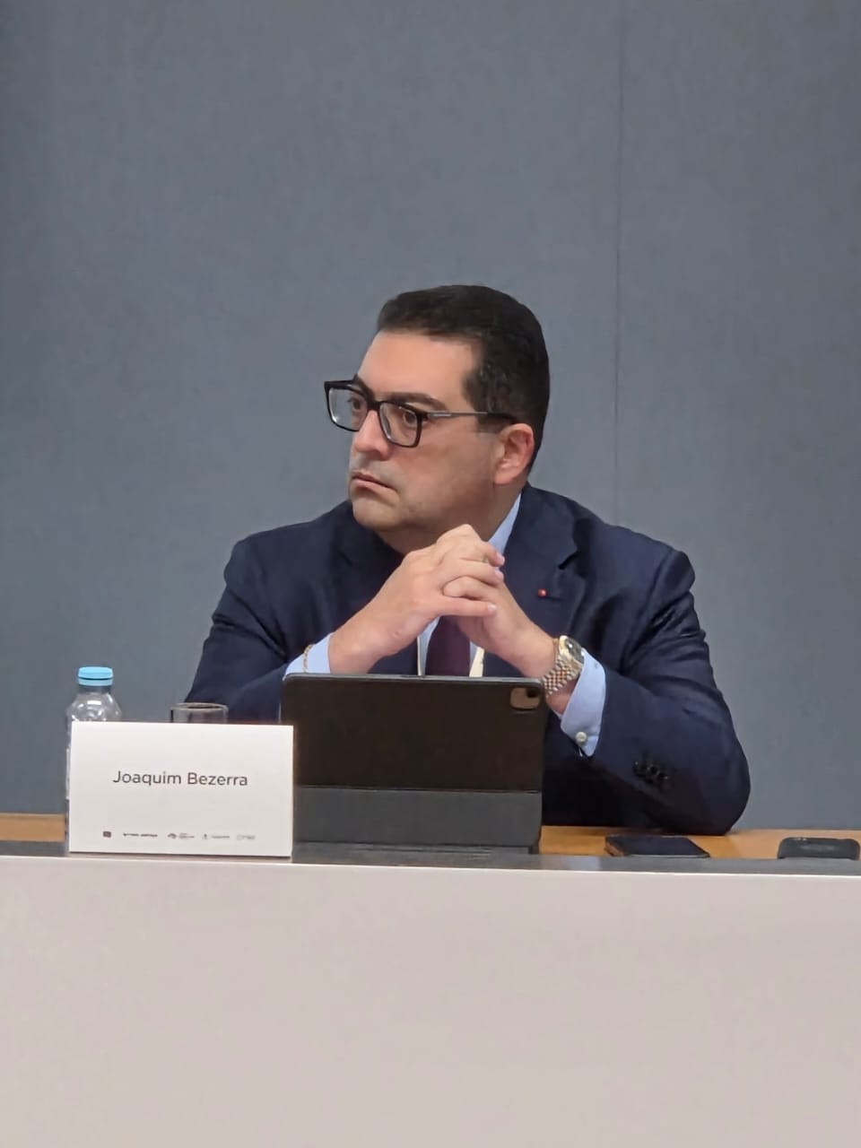 Homem de óculos, terno azul-marinho e gravata vinho, sentado à mesa com as mãos entrelaçadas, olhando para a esquerda. À frente, uma placa com "Joaquim Bezerra" e um tablet.