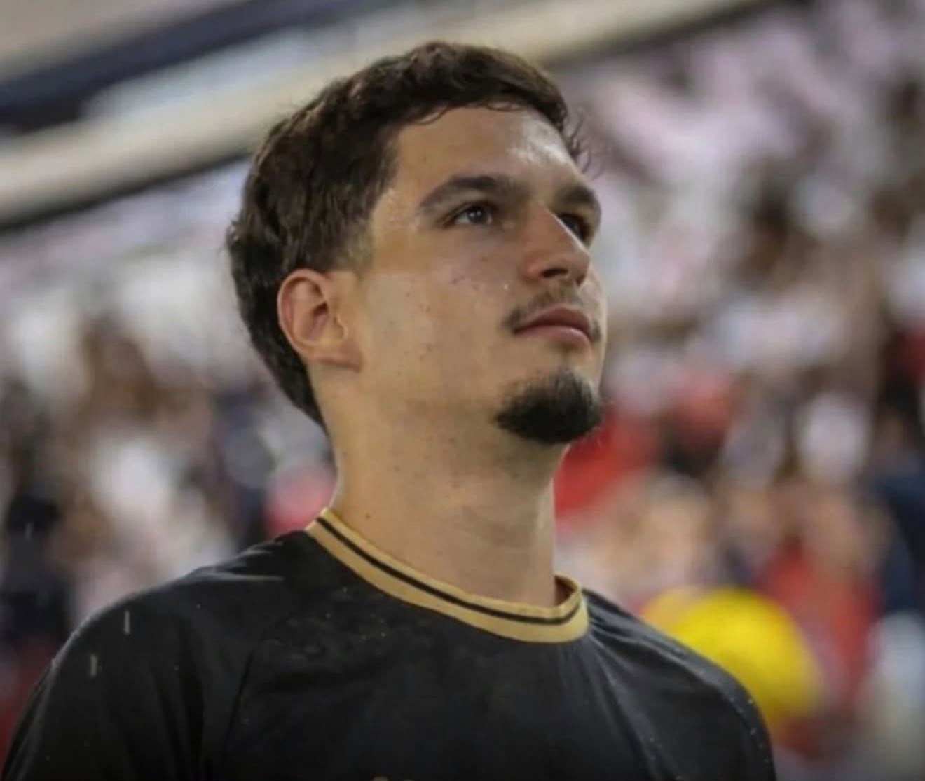 Homem jovem, pele clara, cabelo castanho escuro e barba rala, veste camisa preta com gola dourada, olhando para cima com expressão séria em um estádio
