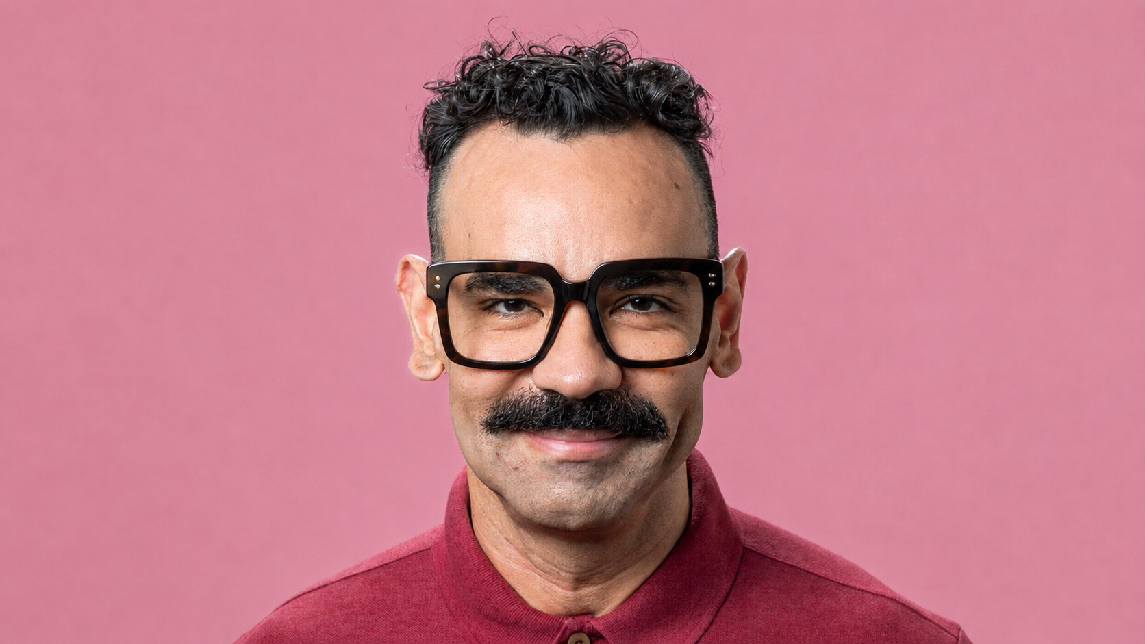Homem de pele morena, bigode preto volumoso e cabelo encaracolado, vestindo camisa polo vinho e óculos de armação quadrada preta, sorrindo para a câmera em fundo rosa