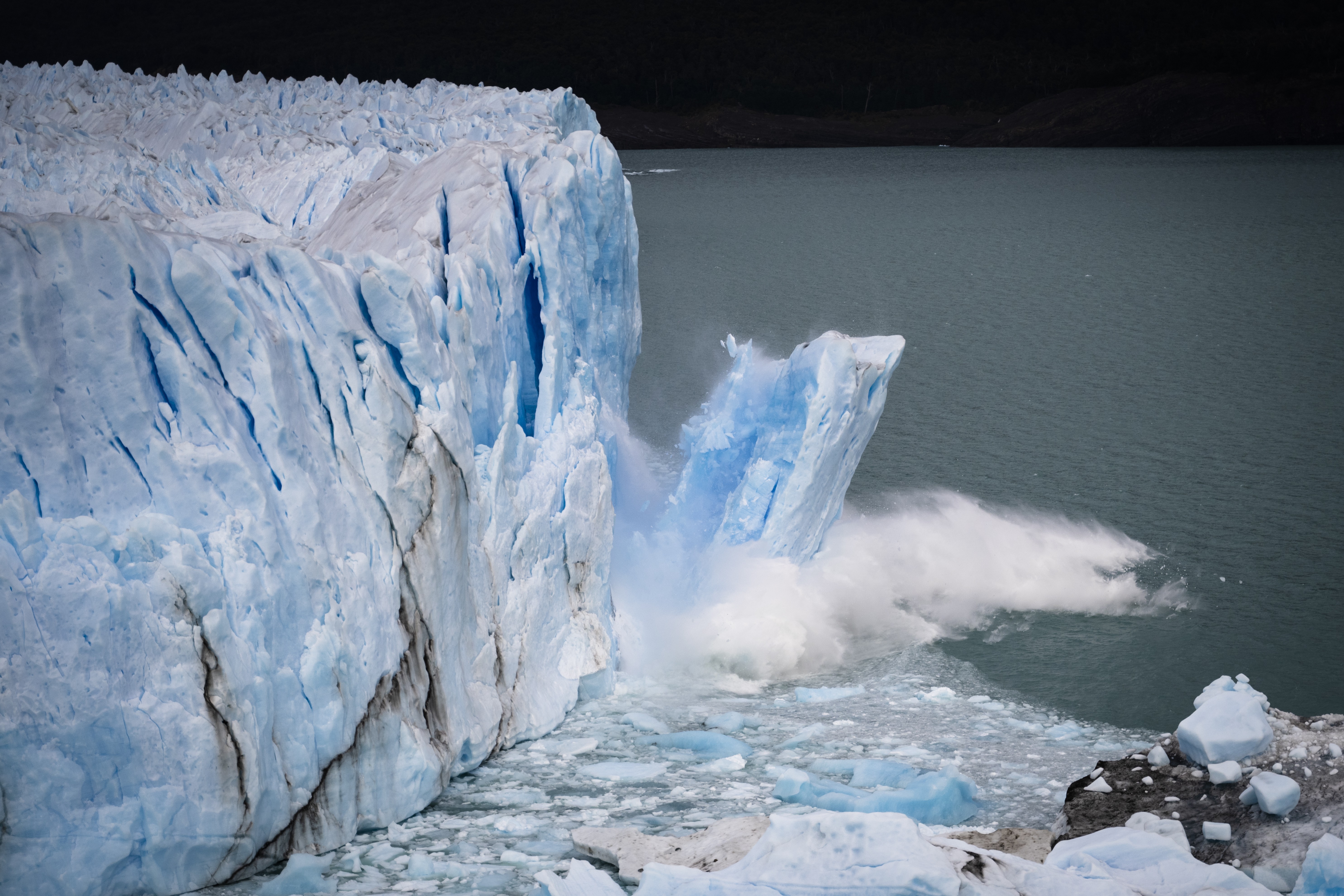Um grande bloco de gelo azul-claro se desprende de uma geleira imponente, caindo na água escura e levantando um spray branco. Pequenos icebergs flutuam na superfície.