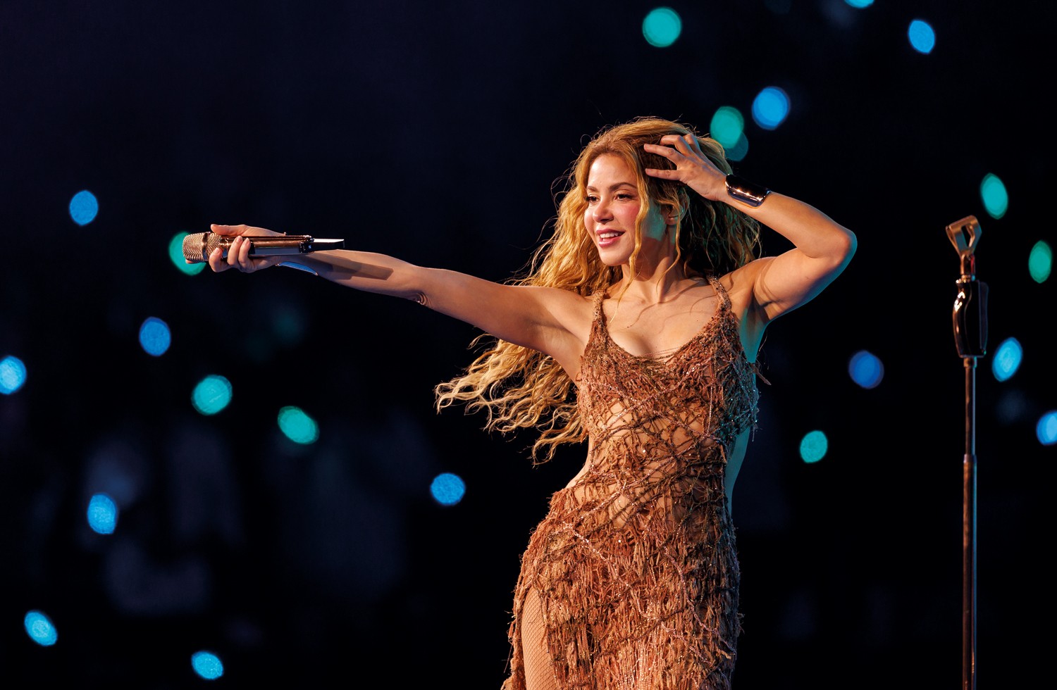 Shakira, com cabelos loiros ondulados, sorri e canta no palco, vestindo um vestido marrom de franjas. Ela segura um microfone dourado com a m&atilde;o direita estendida e a m&atilde;o esquerda na cabe&ccedil;a. O fundo escuro tem luzes azuis e verdes desfocadas