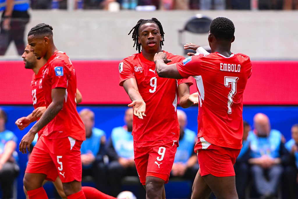 Três jogadores de futebol com uniformes vermelhos em campo. Um jogador com dreads e camisa 9 gesticula com as mãos abertas, enquanto outro, de costas, com camisa 7 e nome EMBOLO, toca seu ombro. Um terceiro jogador, de perfil, com camisa 5, caminha à esquerda.