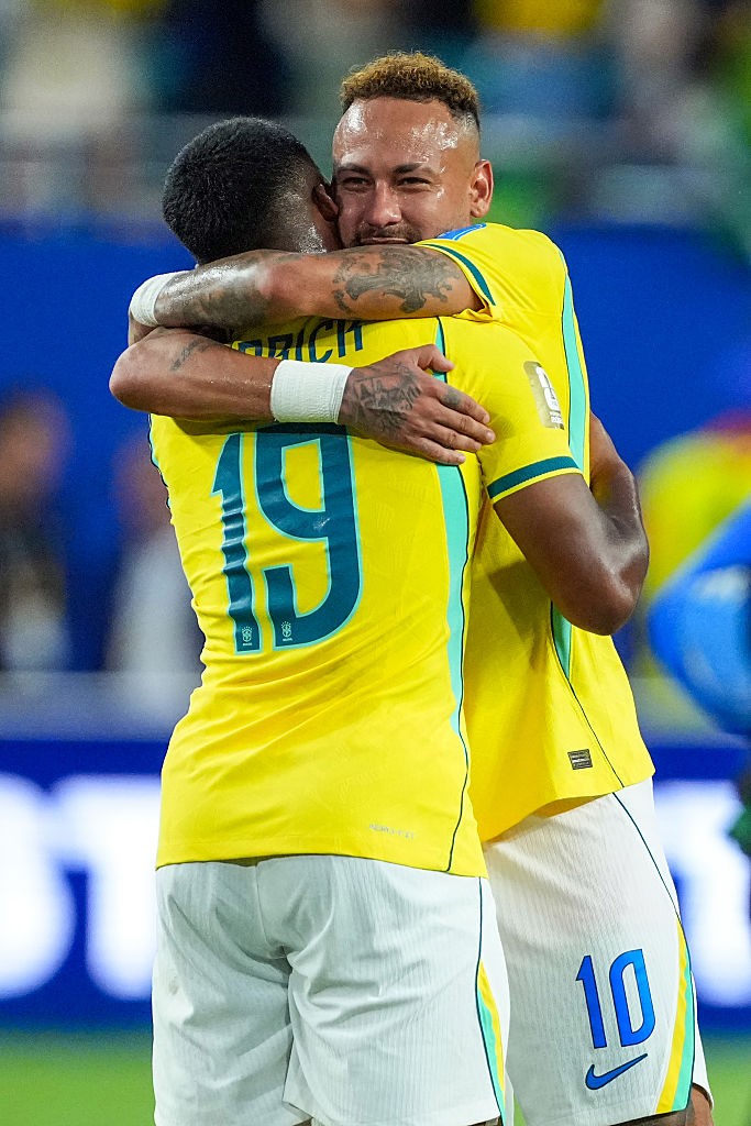 Dois jogadores de futebol brasileiros, um de costas com a camisa 19 e outro de frente com a camisa 10, se abra&ccedil;am no campo, ambos sorrindo e comemorando