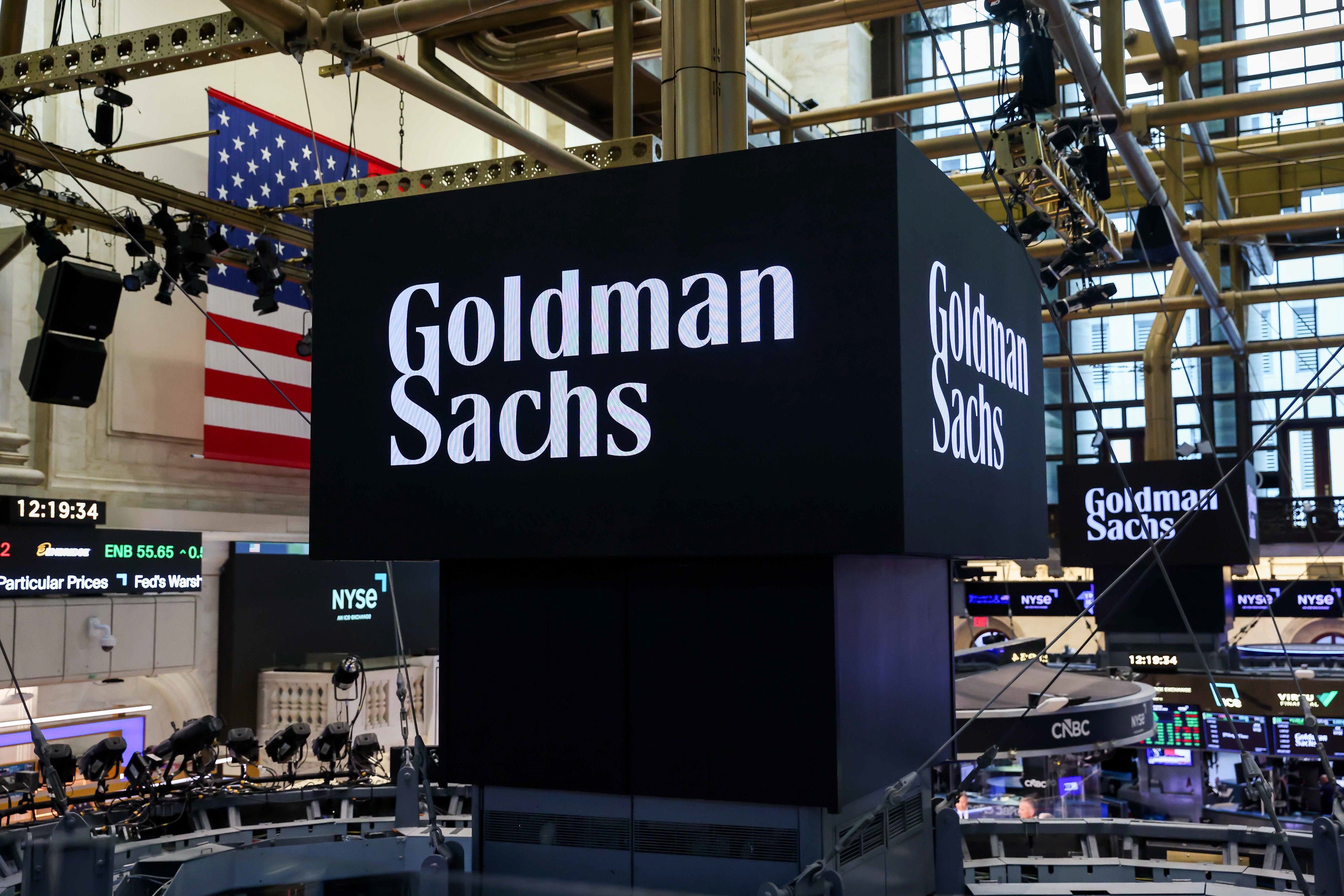 Letreiros digitais pretos com o nome Goldman Sachs em branco, exibidos em um ambiente de bolsa de valores com uma bandeira dos EUA ao fundo e telas de cotações
