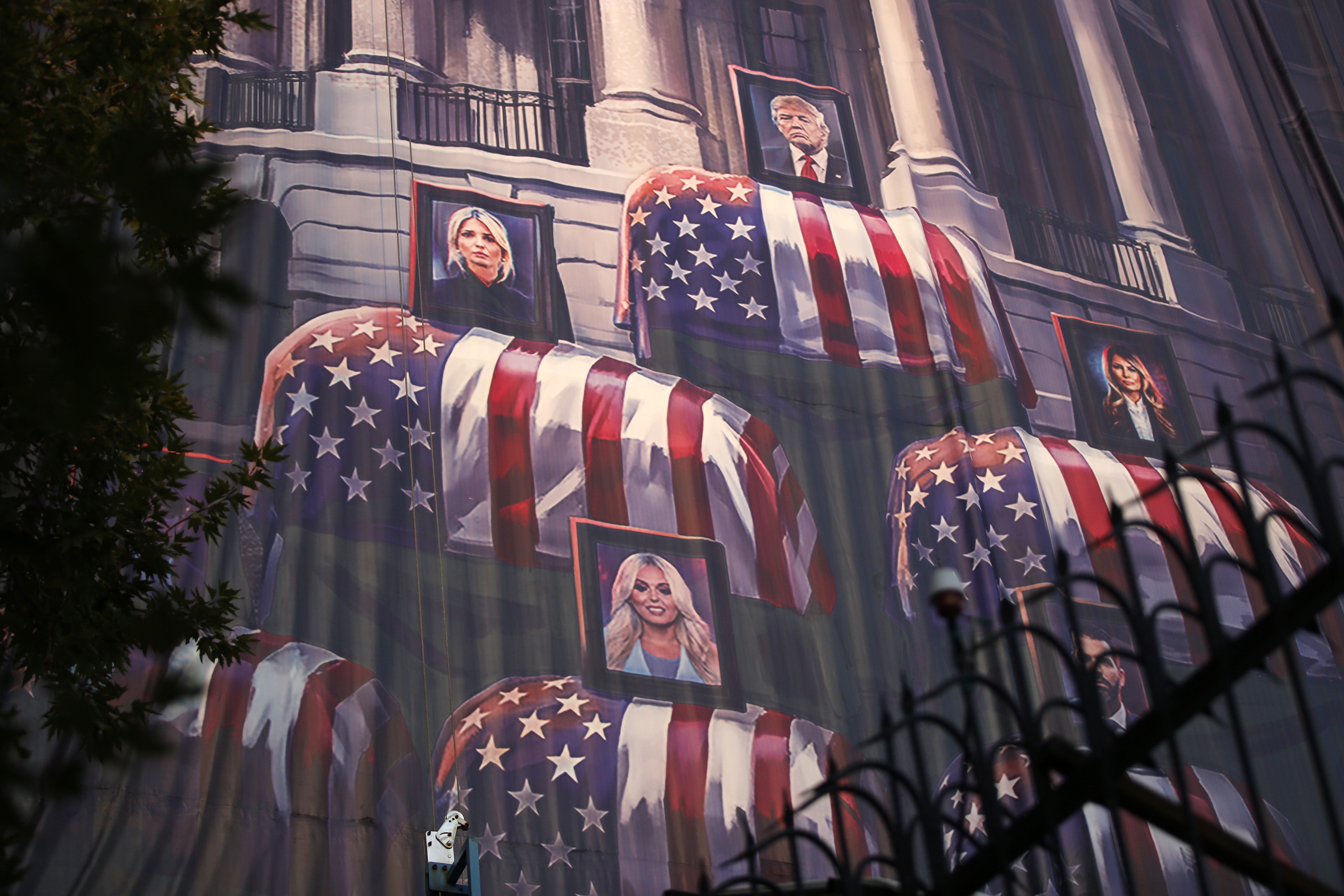Projeção noturna em um prédio com imagens de caixões cobertos por bandeiras dos EUA e retratos de Donald Trump, Melania Trump, Ivanka Trump e outras mulheres loiras. Uma árvore escura e um portão de ferro forjado aparecem em primeiro plano