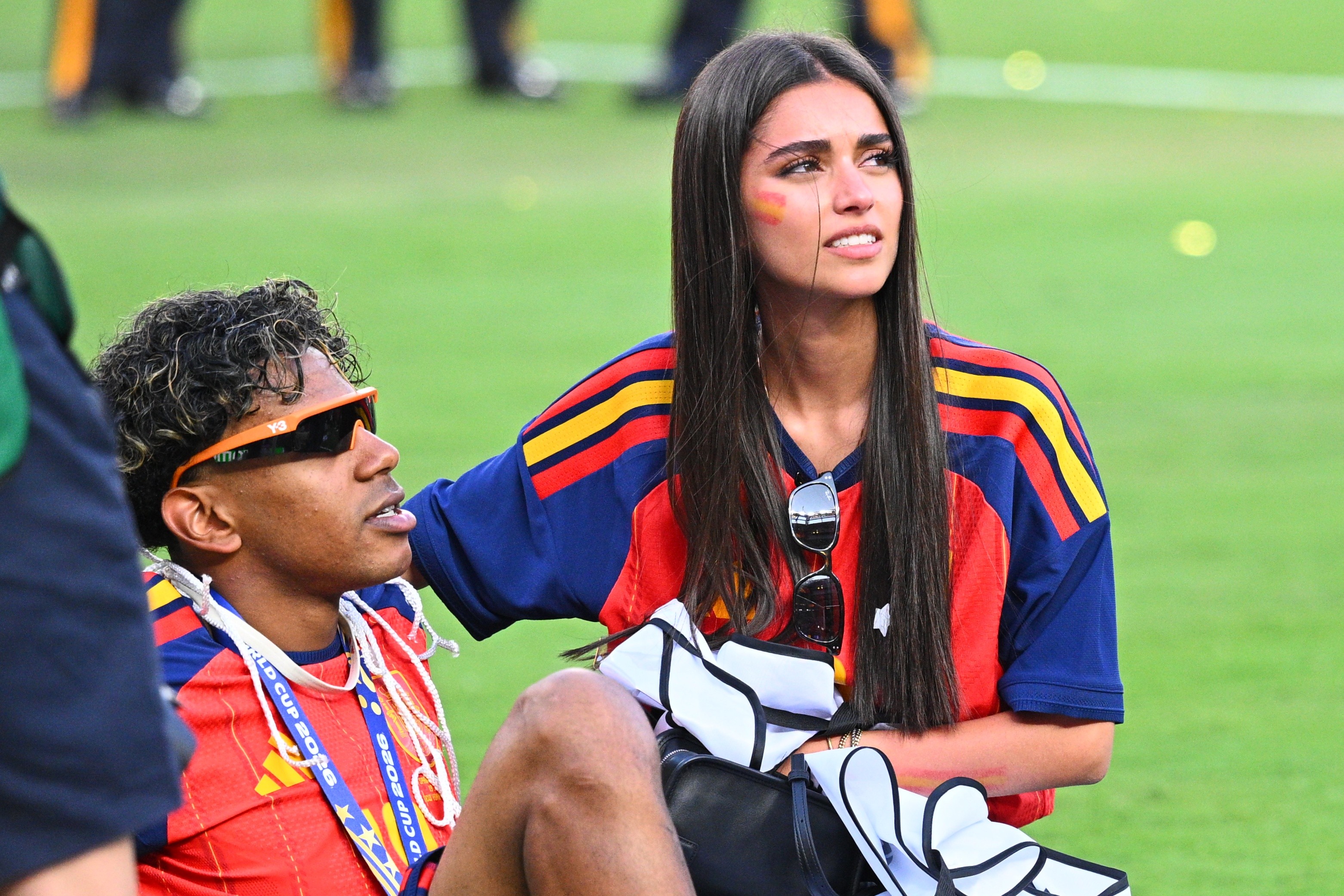 Um homem e uma mulher sentados em um campo de grama verde. O homem, à esquerda, usa óculos de sol laranja e uma camisa vermelha com cordões azuis. A mulher, à direita, tem cabelos longos e escuros, usa uma camisa azul e vermelha com listras amarelas, óculos de sol pendurados na gola e uma pintura facial de bandeira no rosto. Ambos olham para a direita, com expressões atentas
