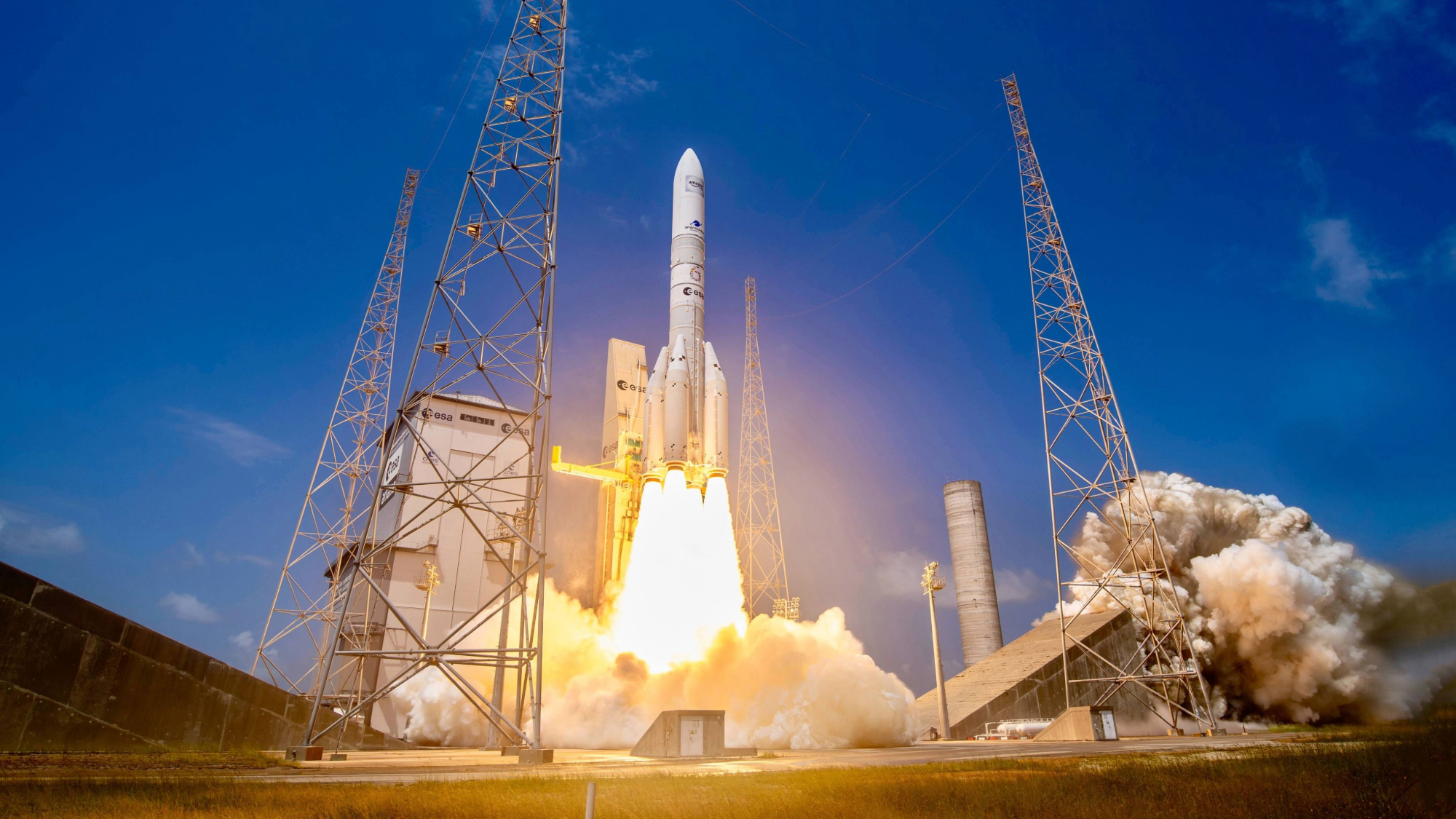 Foguete branco Ariane 5 decolando, com chamas e fumaça densa saindo da base, em uma plataforma de lançamento com torres metálicas e céu azul