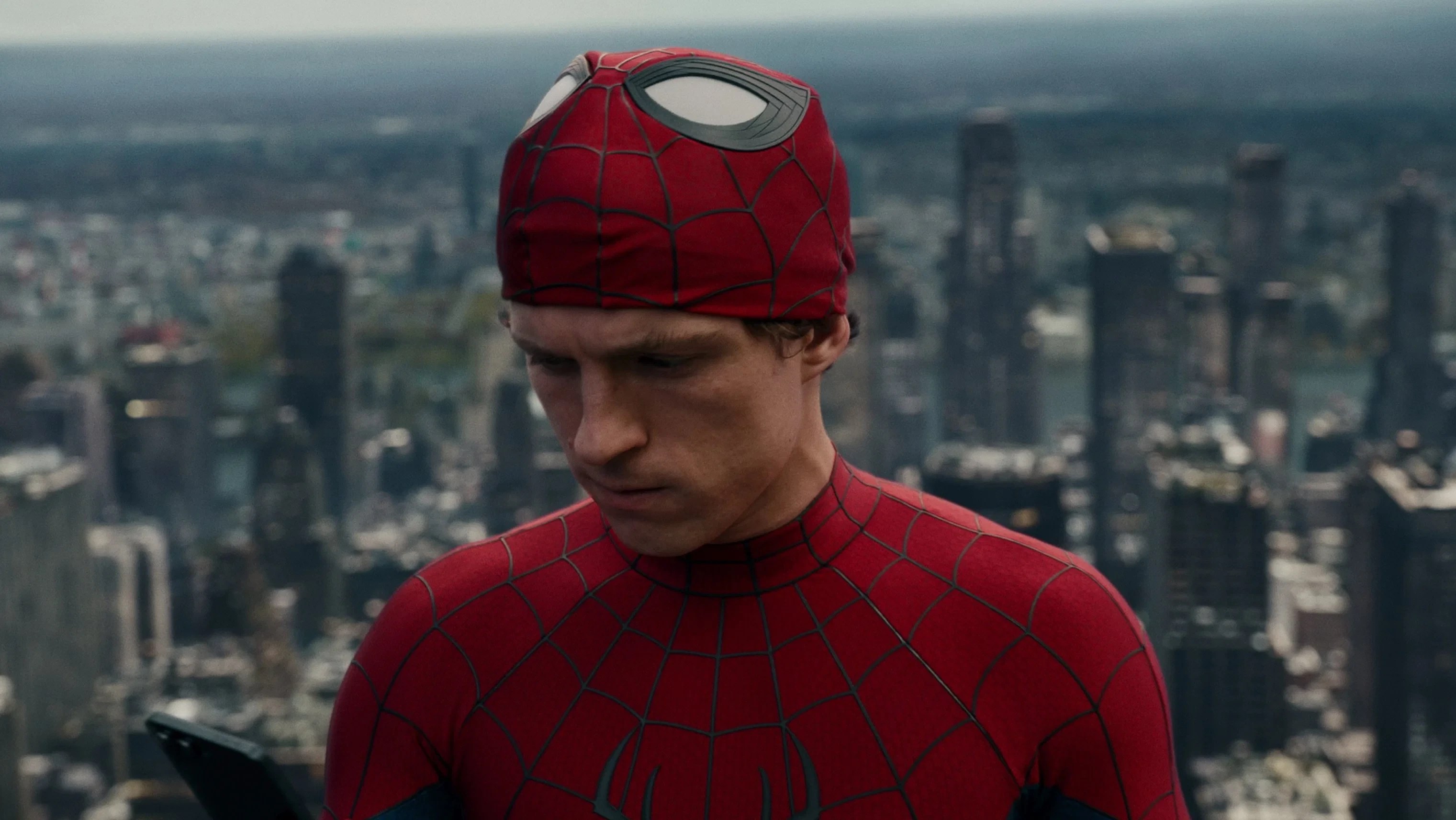 Homem-Aranha, com parte da máscara levantada revelando seu rosto sério, olha para baixo enquanto segura um celular. Ele veste o traje vermelho e azul, com a cidade ao fundo