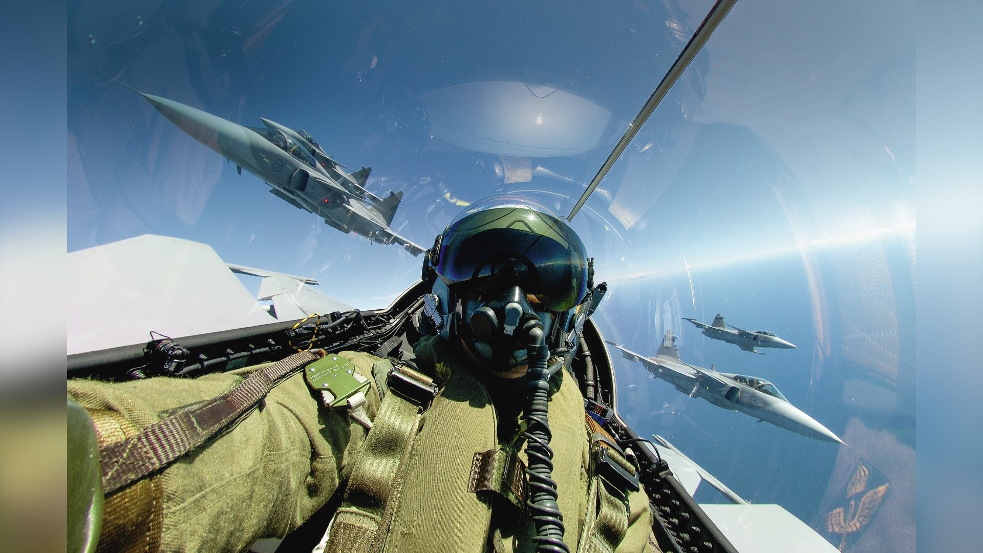 Piloto de caça em uniforme verde e capacete com viseira escura, dentro da cabine de um avião, com três jatos militares voando ao redor sob um céu azul claro