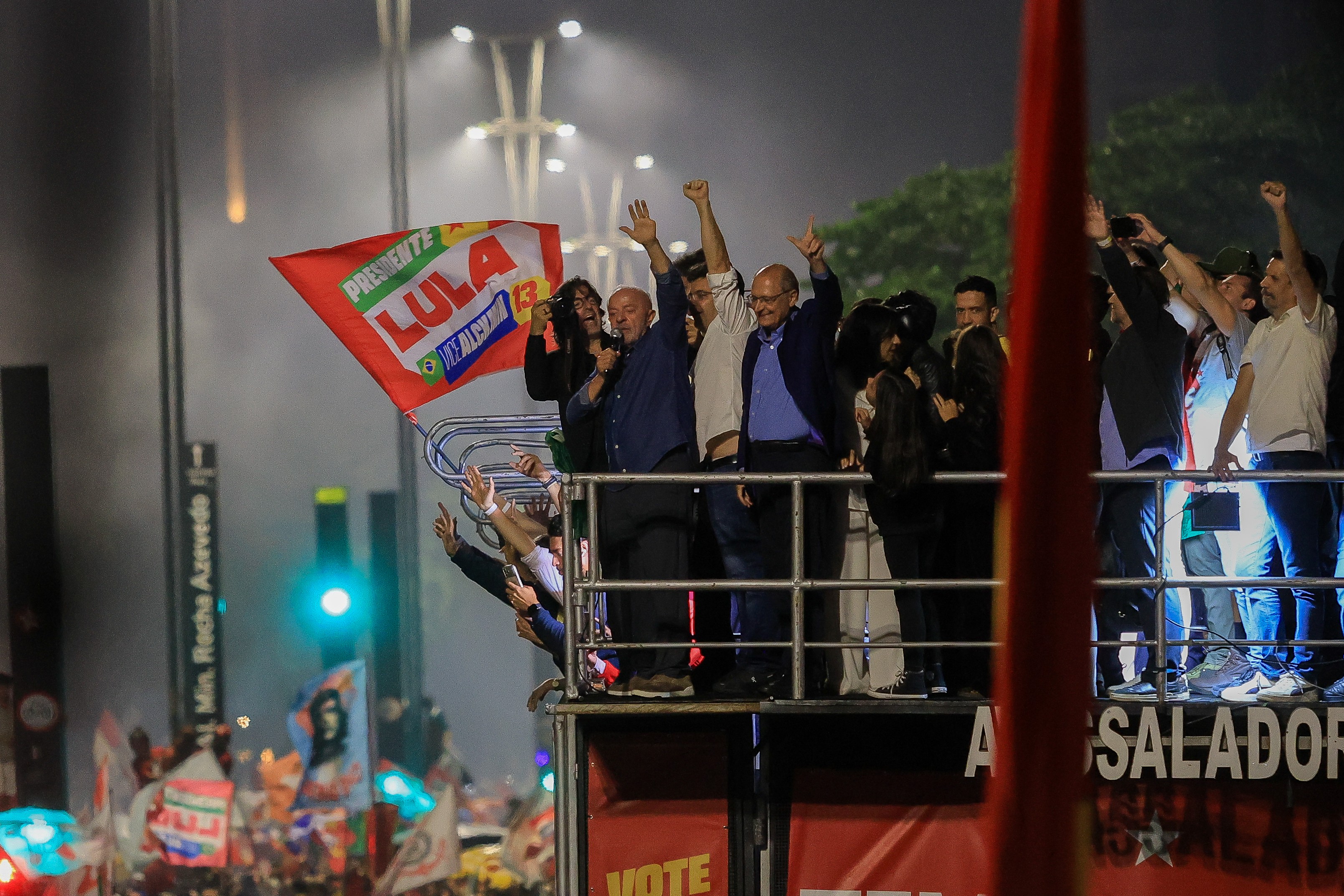 Lula e Geraldo Alckmin acenam para a multidão de cima de um trio elétrico à noite, com uma bandeira vermelha LULA PRESIDENTE 13 ao lado.