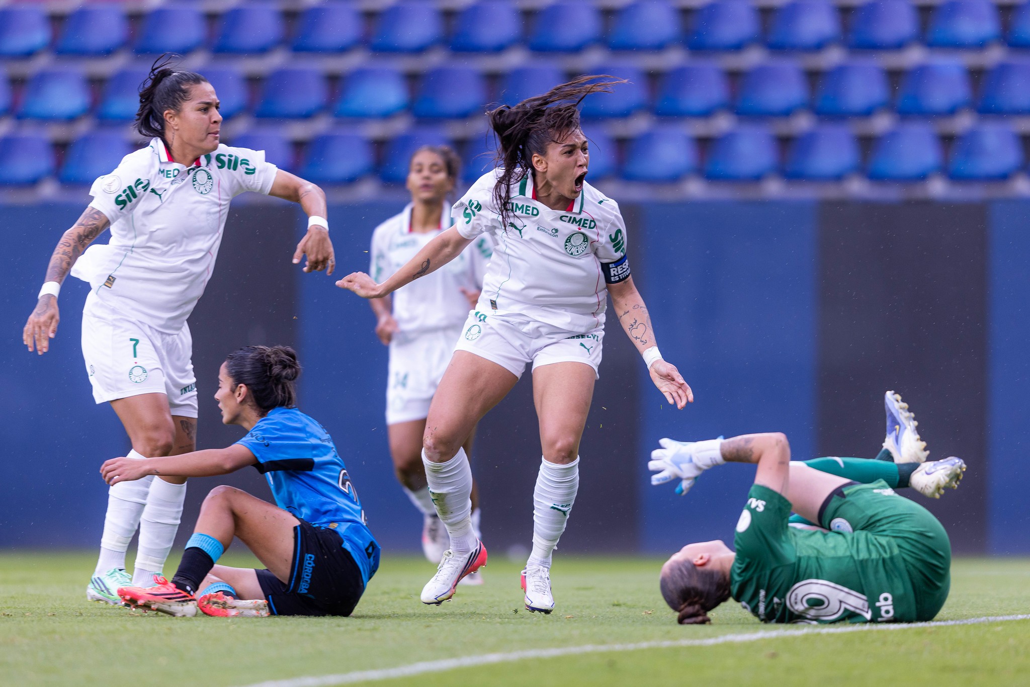 Jogadoras de futebol feminino em campo. Uma atleta de camisa branca e shorts brancos, com cabelo preso, grita com a boca aberta e braços flexionados. Outra jogadora de camisa branca e shorts brancos, com cabelo preso, observa. Uma terceira, de camisa azul e shorts pretos, está sentada no chão. Uma goleira de camisa verde e luvas brancas está deitada de costas no gramado. Cadeiras azuis vazias ao fundo