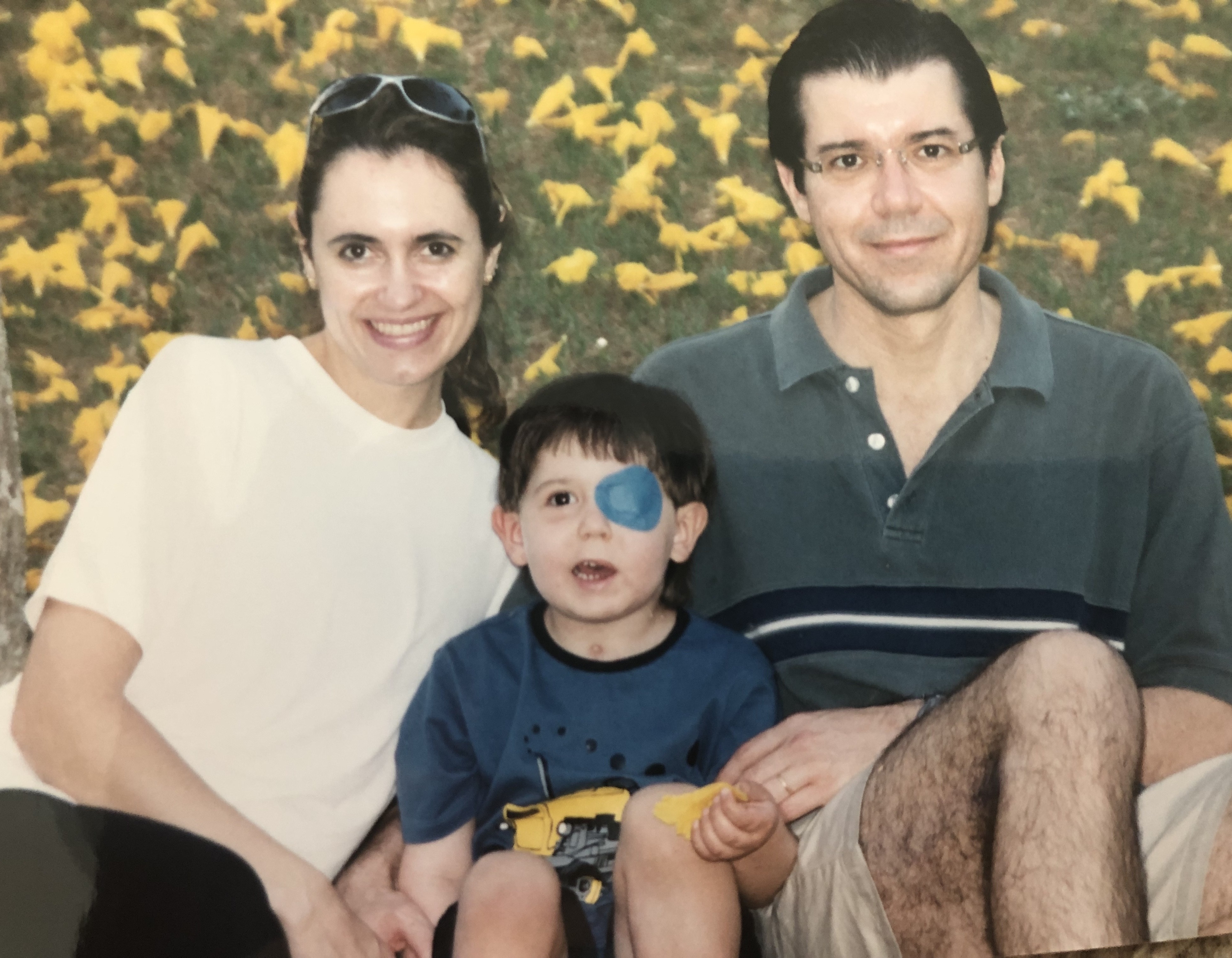 Família sorridente com um menino pequeno usando um tapa-olho azul, sentados em frente a uma parede de folhas amarelas