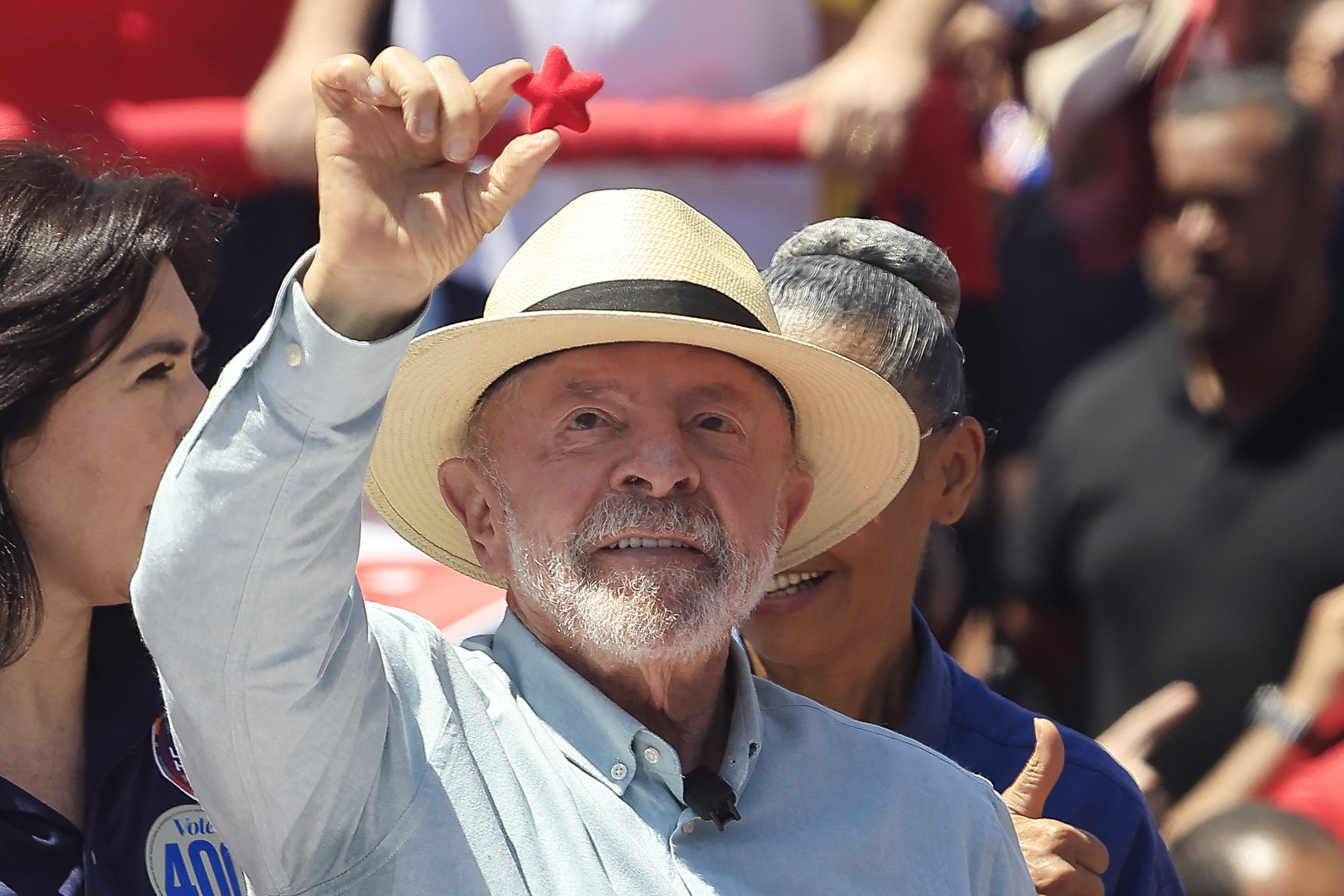 Homem idoso de barba branca e chapéu de palha claro, sorrindo, ergue a mão direita segurando uma estrela vermelha. Ao seu lado, uma mulher de cabelo escuro e um homem sorridente.