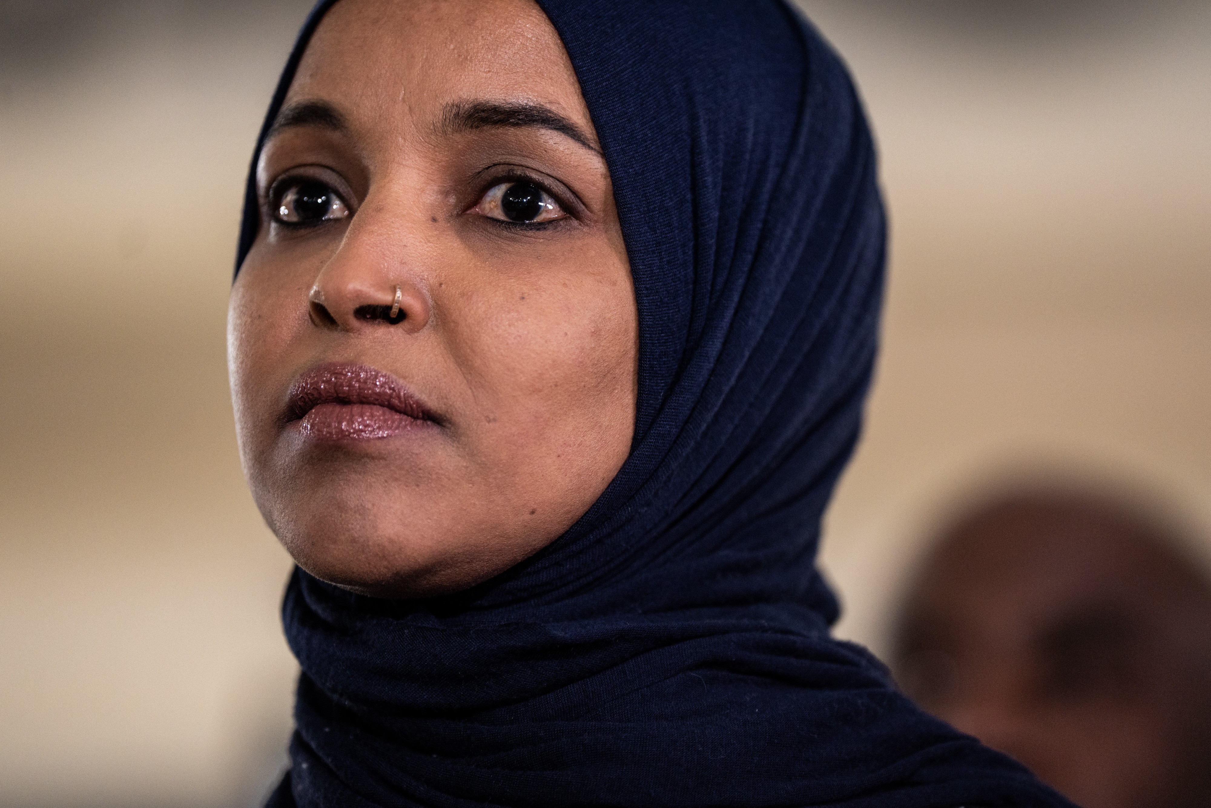 Mulher de pele morena, com um hijab azul-marinho e um piercing dourado no nariz, olha para cima com expressão séria. Seus lábios têm um batom brilhante e seus olhos estão delineados. O fundo está desfocado