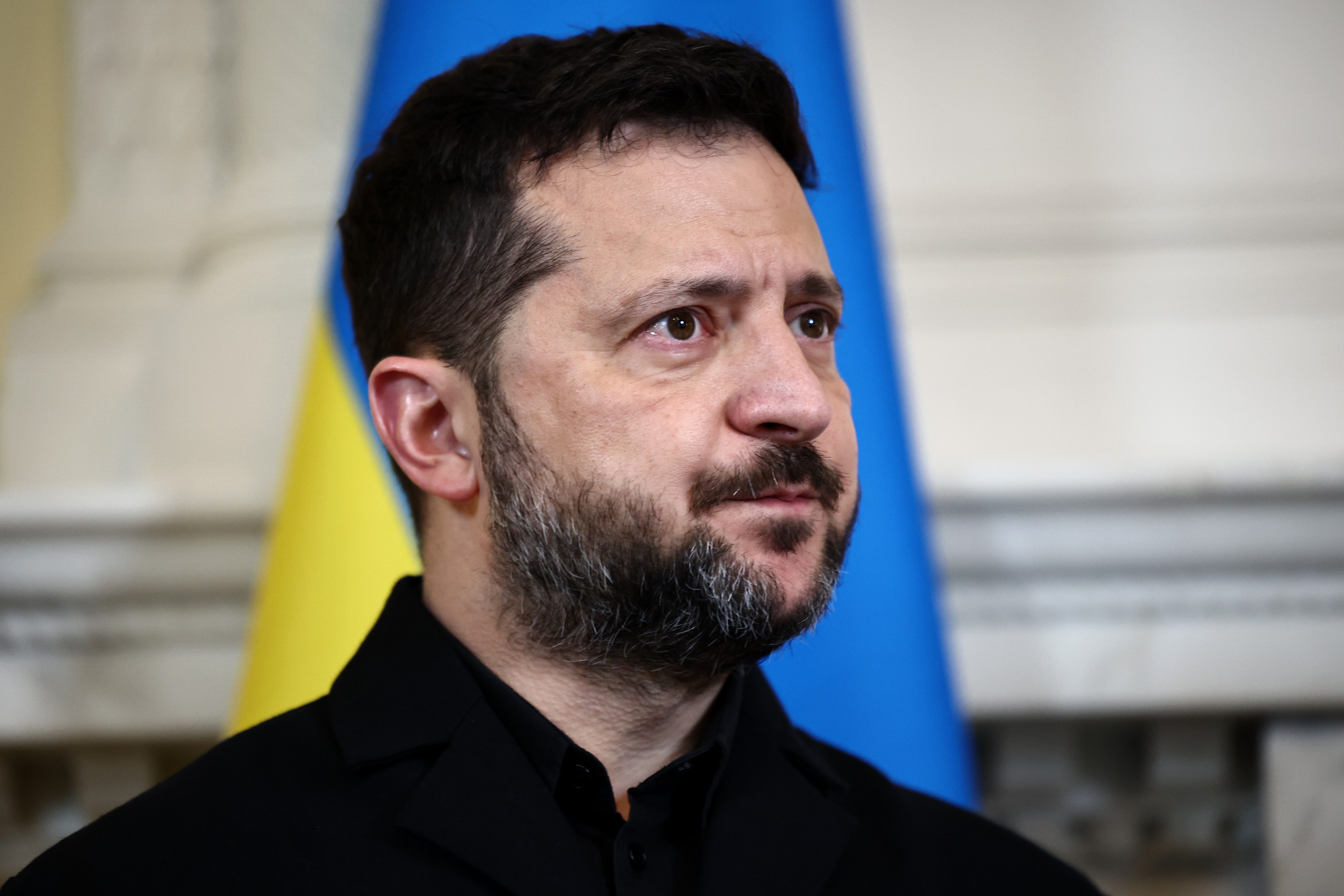 Volodymyr Zelensky, presidente da Ucrânia, com barba escura e cabelo curto, veste uma camisa preta. Ele olha para a direita, com a bandeira ucraniana azul e amarela desfocada ao fundo