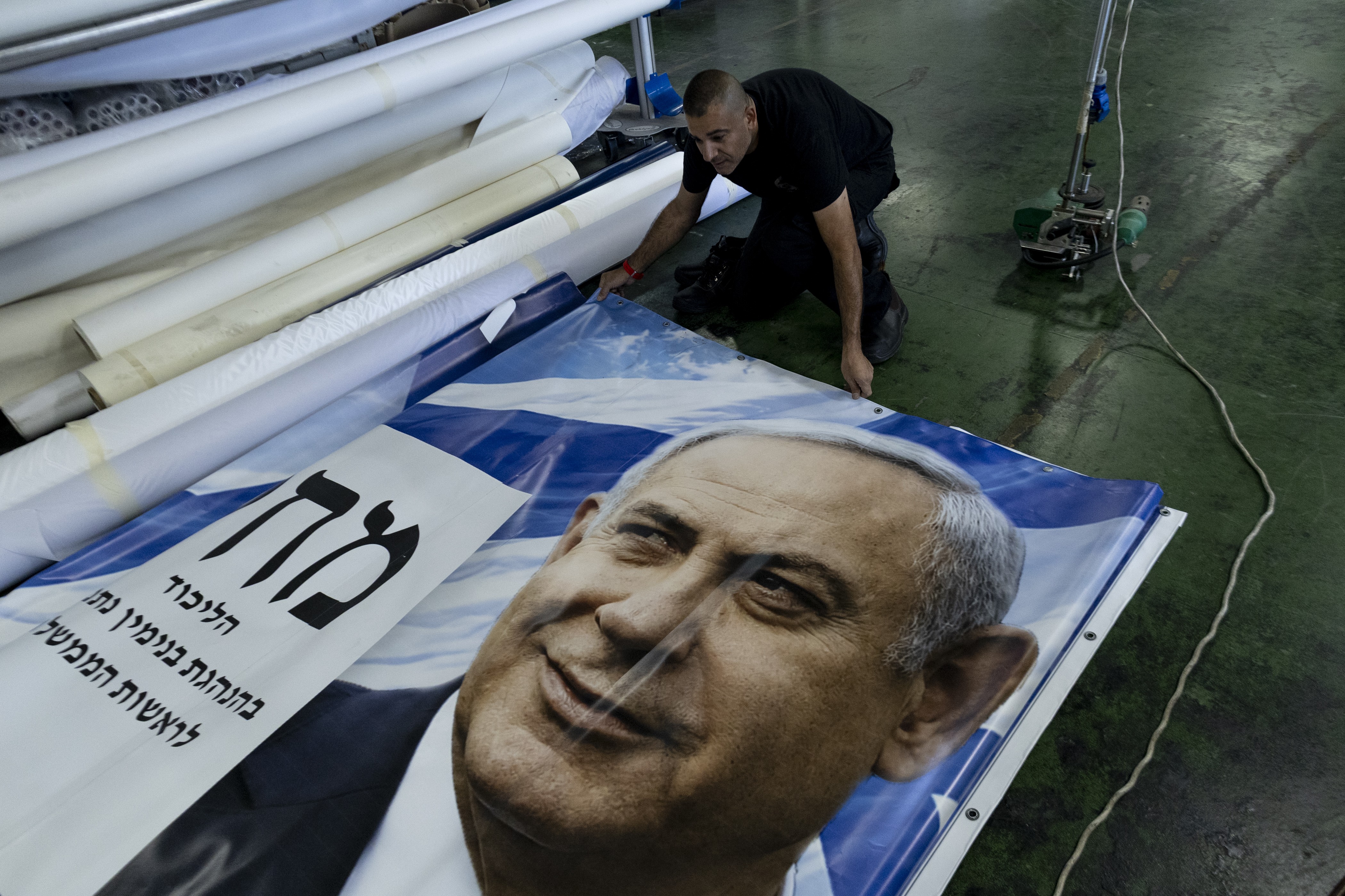 Homem de camiseta preta e calça escura ajoelhado, desdobrando um grande banner azul e branco com o rosto sorridente de Benjamin Netanyahu e texto em hebraico. Rolos de tecido branco estão empilhados ao fundo, e um cabo elétrico verde se estende pelo chão.